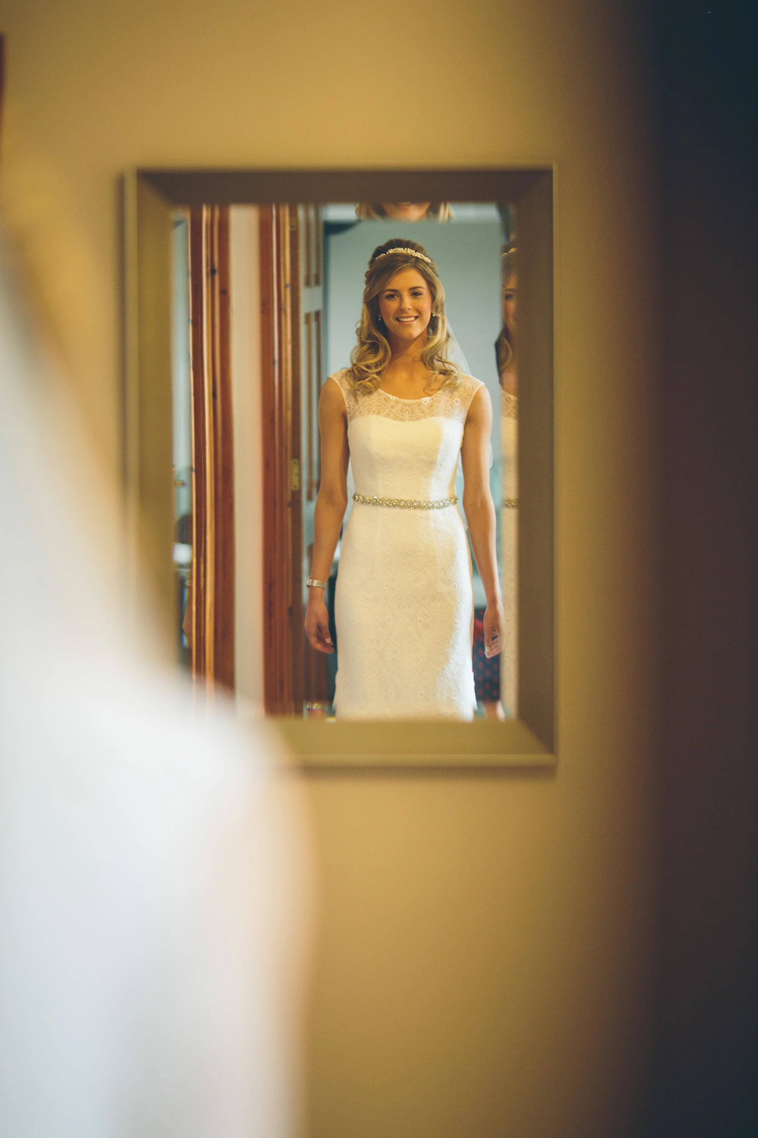 A bride in a white lace wedding dress with a belt stands in front of a mirror, smiling.