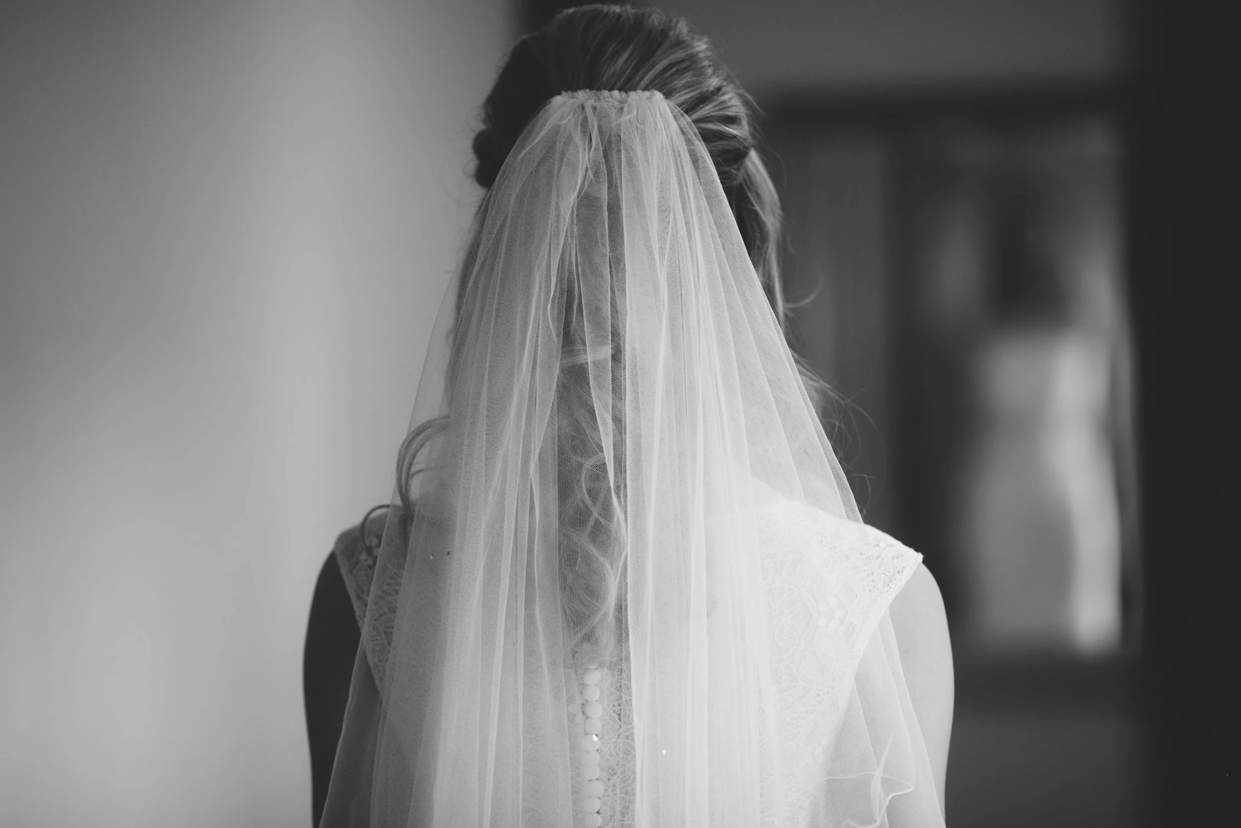 A black and white photo of a bride with her face hidden behind a veil, looking towards a mirror.