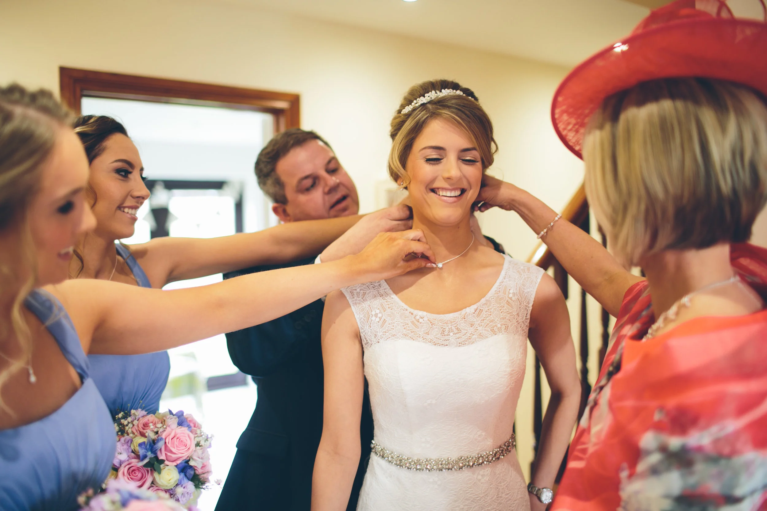 A bride in a white dress is surrounded by her bridesmaids and family as they help her put on a necklace, all smiling and smiling in a brightly lit room.
