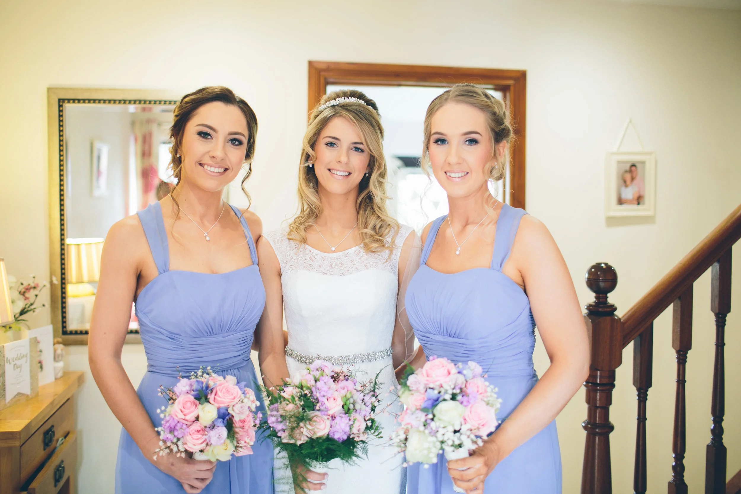 A bride and two bridesmaids standing inside a house, smiling, with the bride in a white wedding dress and the bridesmaids in light purple dresses, holding bouquets of flowers.