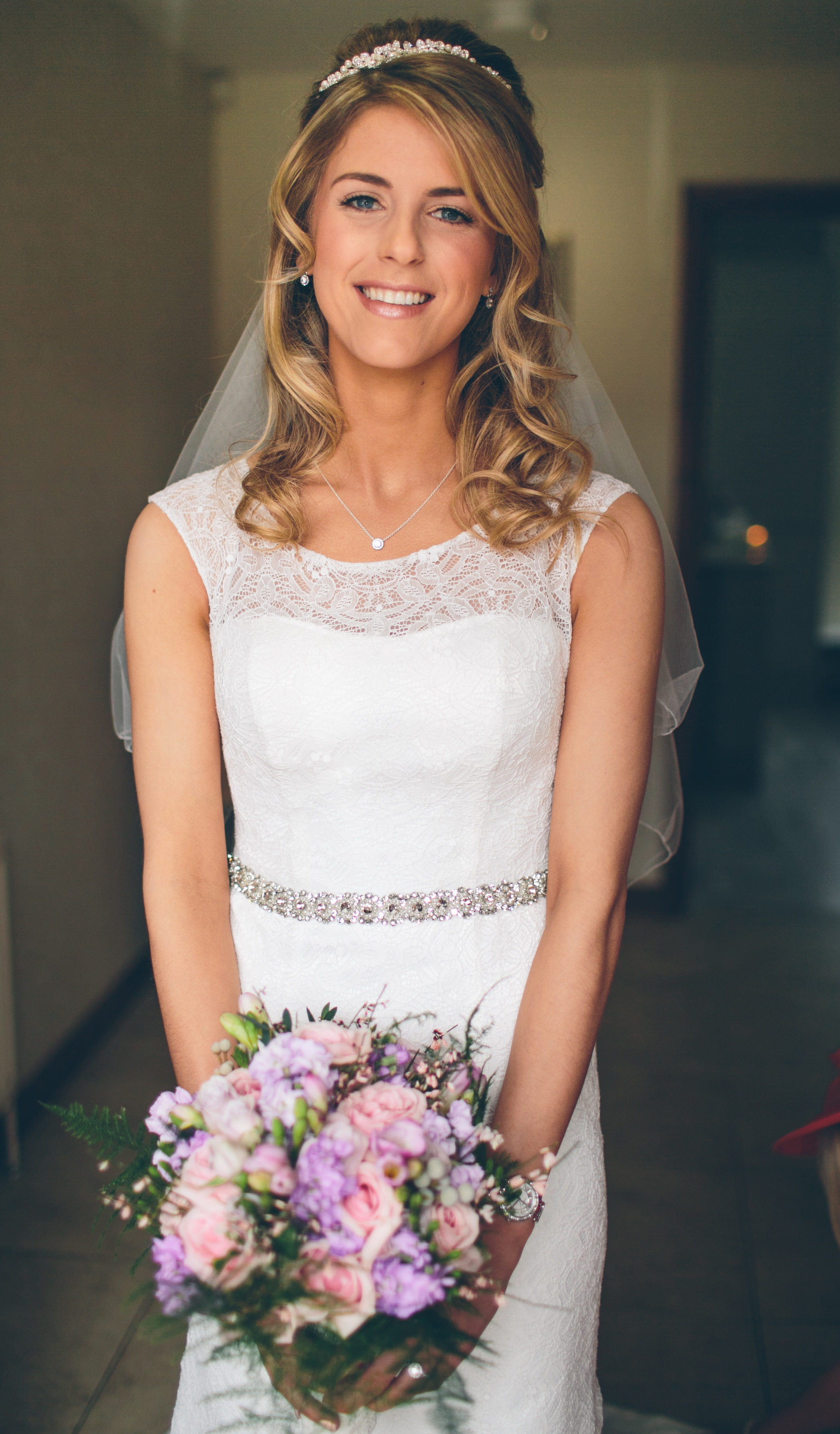A smiling blonde bride in a white lace wedding dress holding a bouquet of pink, purple, and white flowers standing indoors.