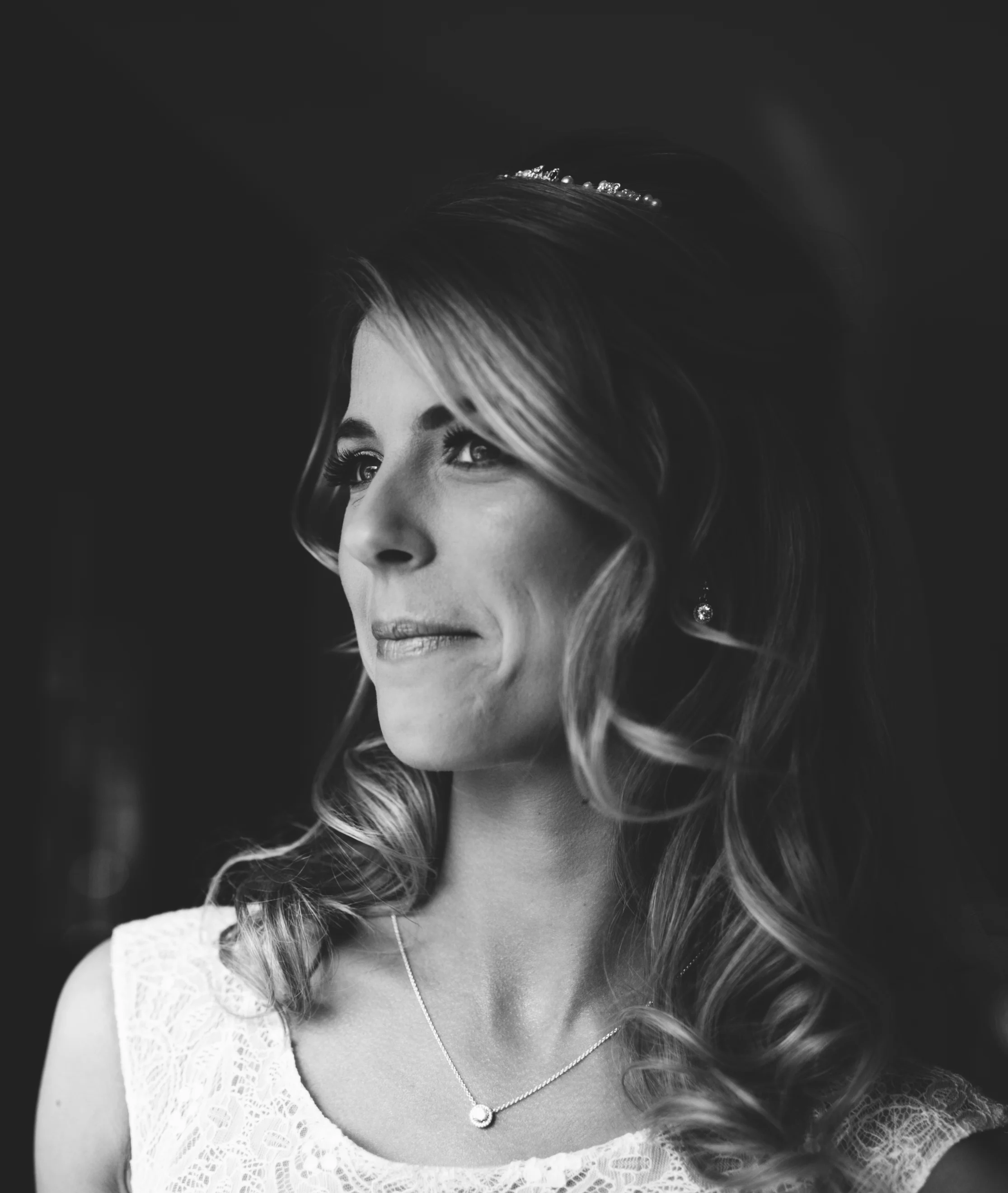Black and white portrait of a woman with wavy hair wearing a lace dress, a necklace, and earrings, looking to the side with a slight smile.