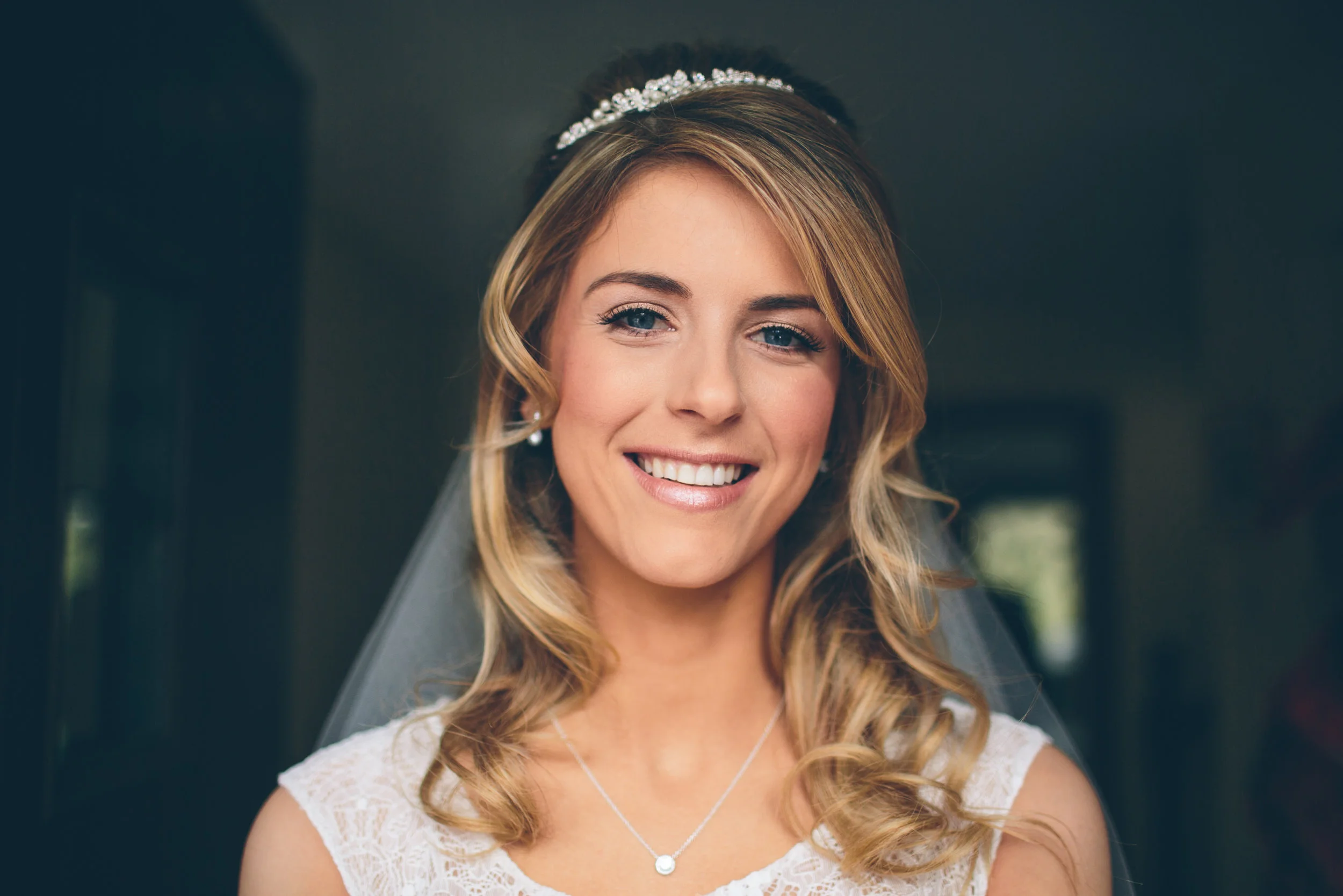 A woman with blonde hair wearing a white lace dress, a pearl tiara, and a necklace, smiling indoors.