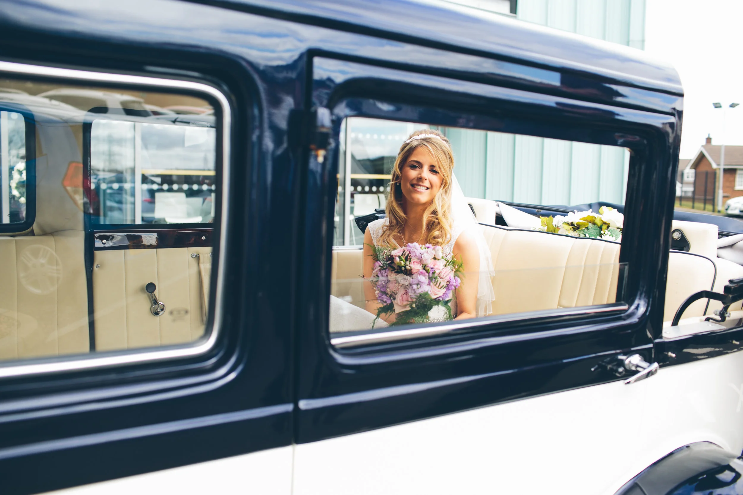 A smiling woman in a wedding dress sitting inside a vintage car, holding a bouquet of pink and purple flowers.