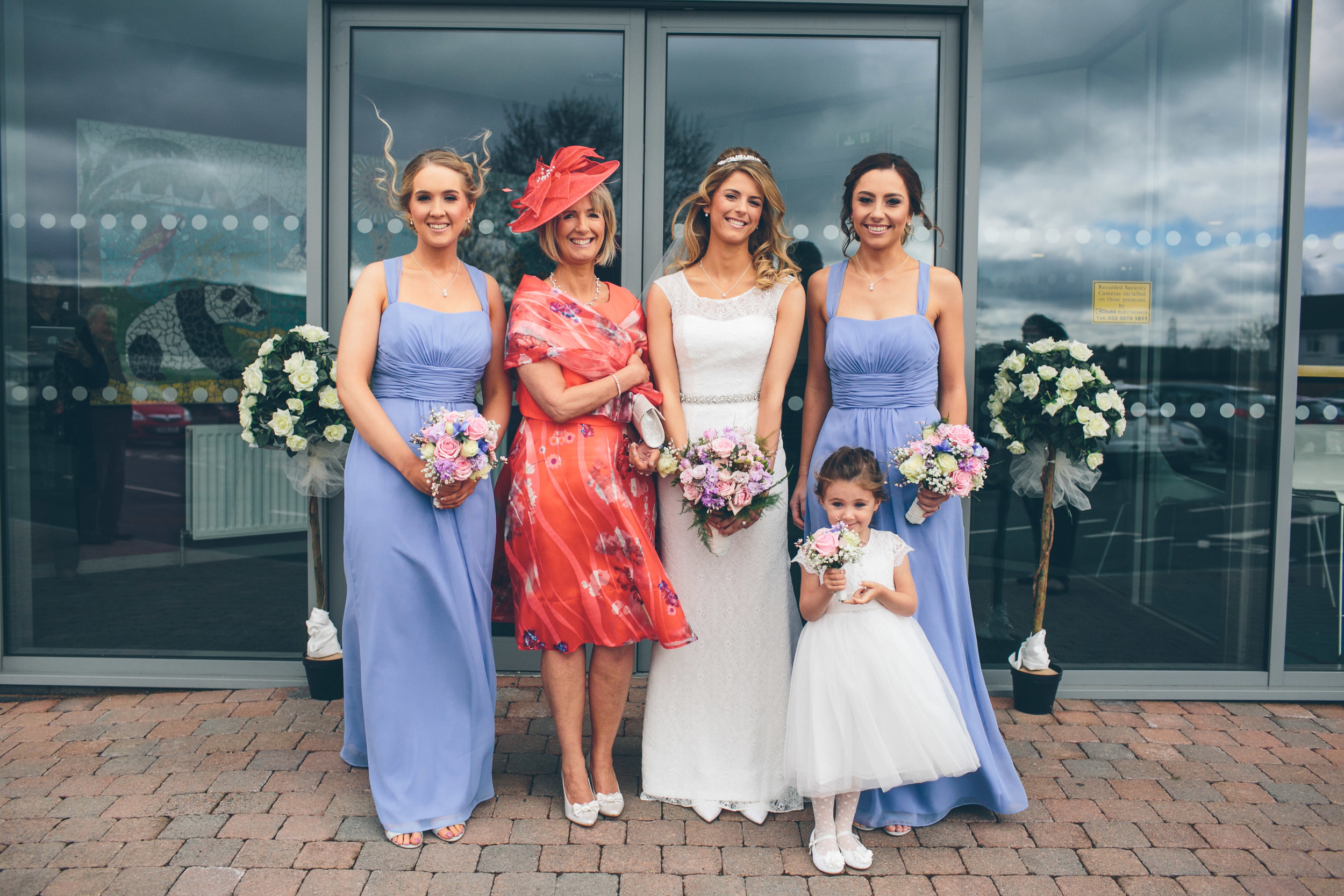 A group of six women and a young girl standing in front of a glass building, at a wedding. The bride is in the middle wearing a white wedding dress, surrounded by bridesmaids in blue dresses. The young girl is in a white dress holding a small bouquet