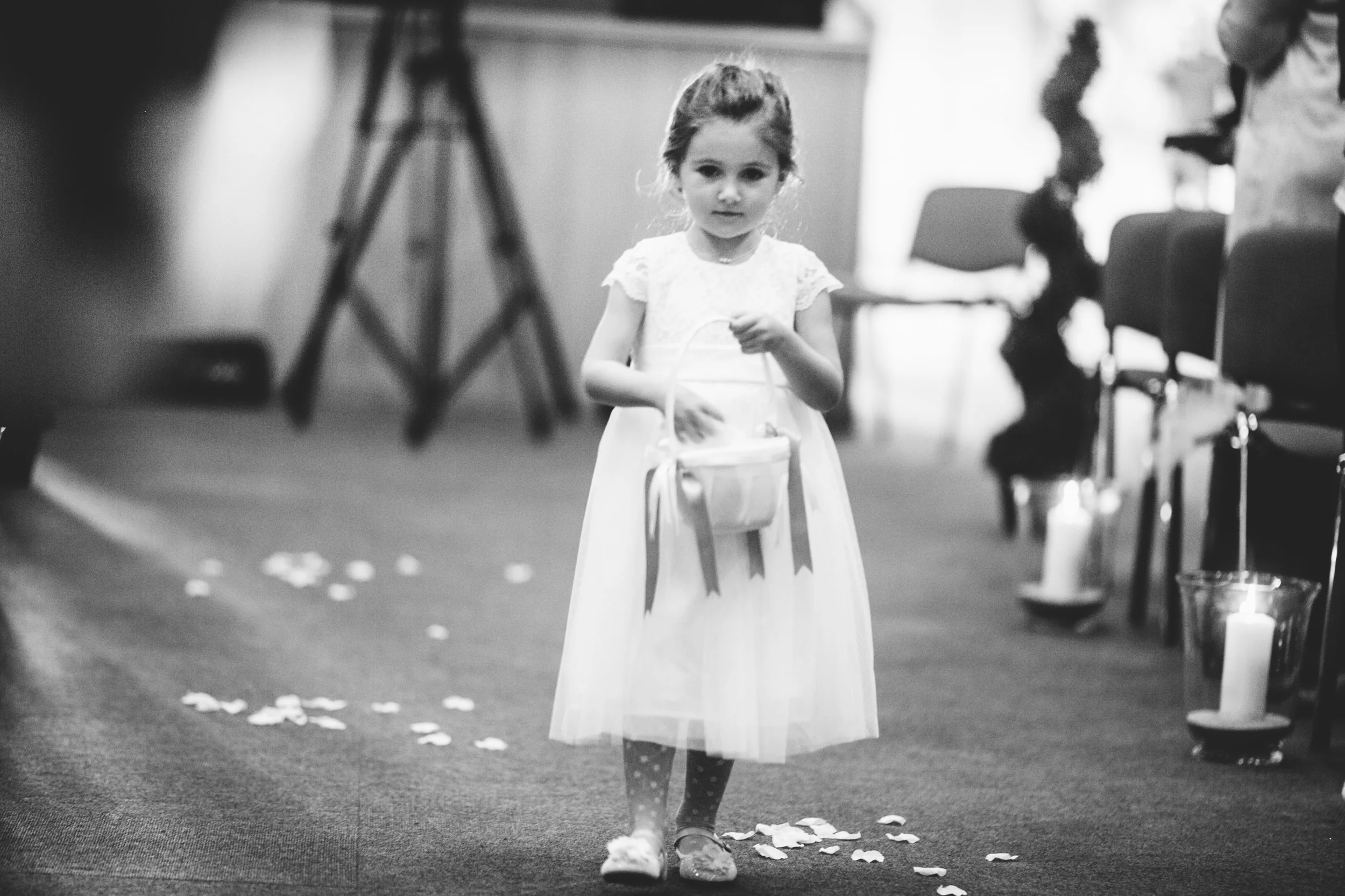 A young girl in a white dress walking down an aisle decorated with scattered flower petals, holding a small basket, at what appears to be a wedding or celebration.
