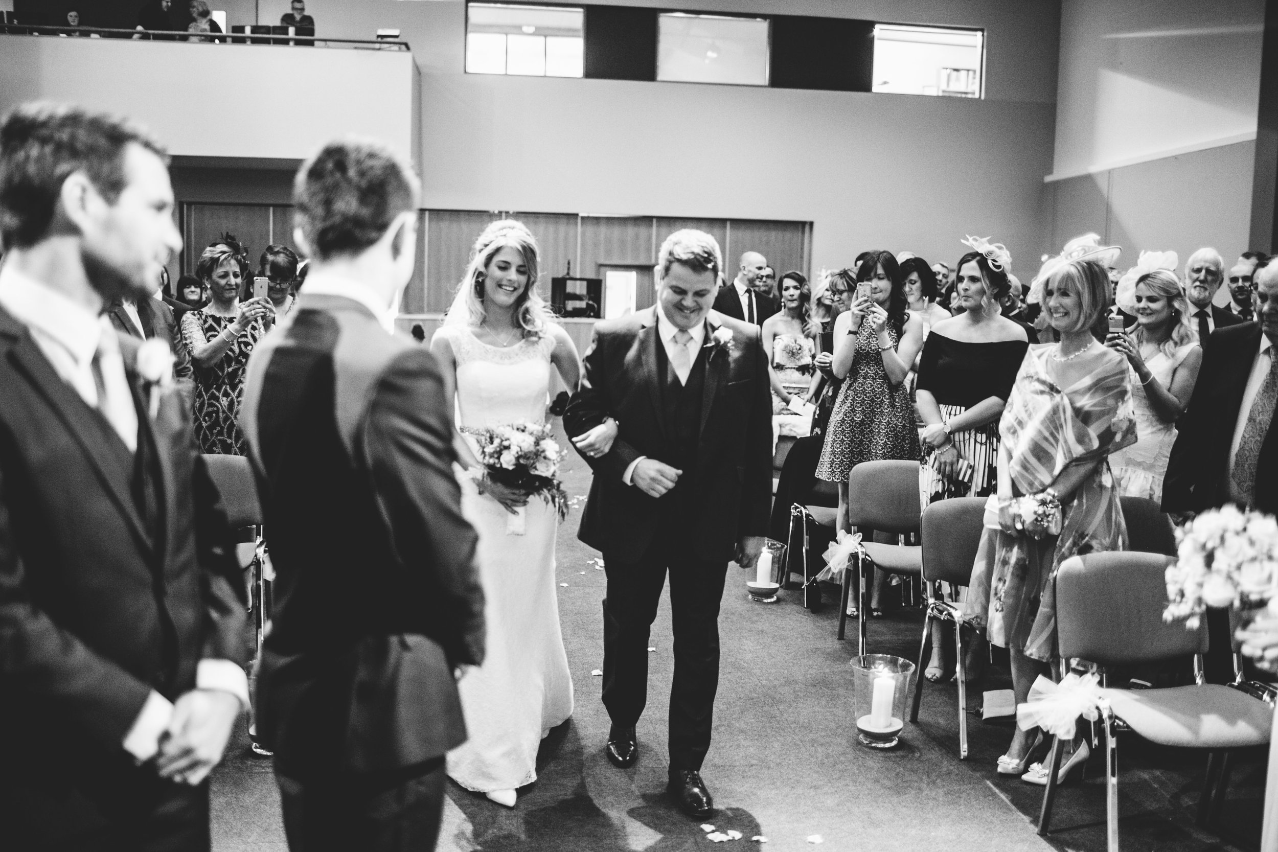 A black-and-white photo of a wedding ceremony with the bride and groom walking down the aisle, surrounded by seated guests who are taking pictures and smiling.