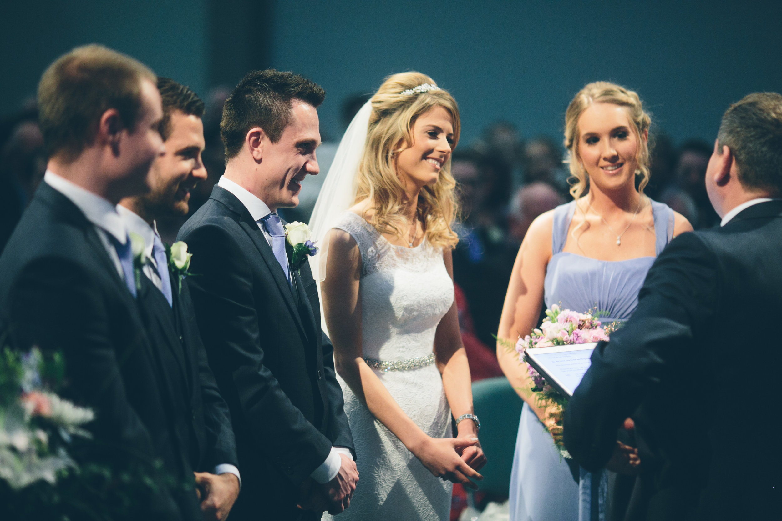 Bride and groom exchanging wedding vows with wedding party and guests in background.