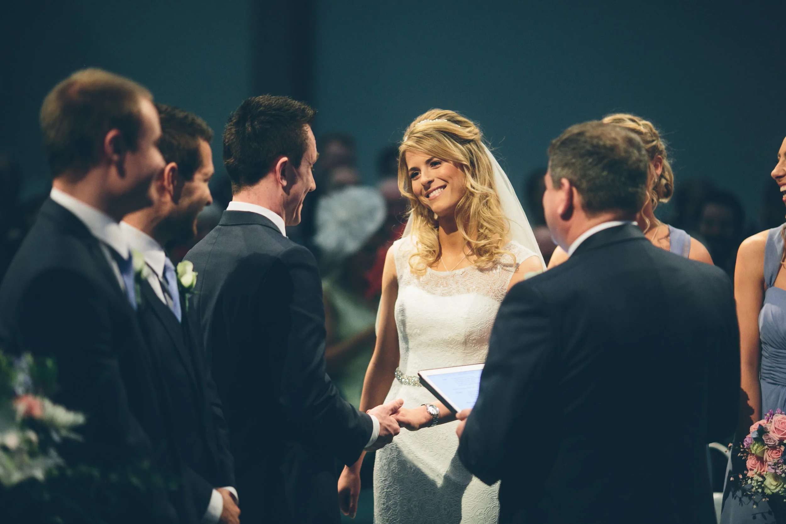 A bride and groom exchange vows at their wedding ceremony, surrounded by groomsmen and bridesmaids.