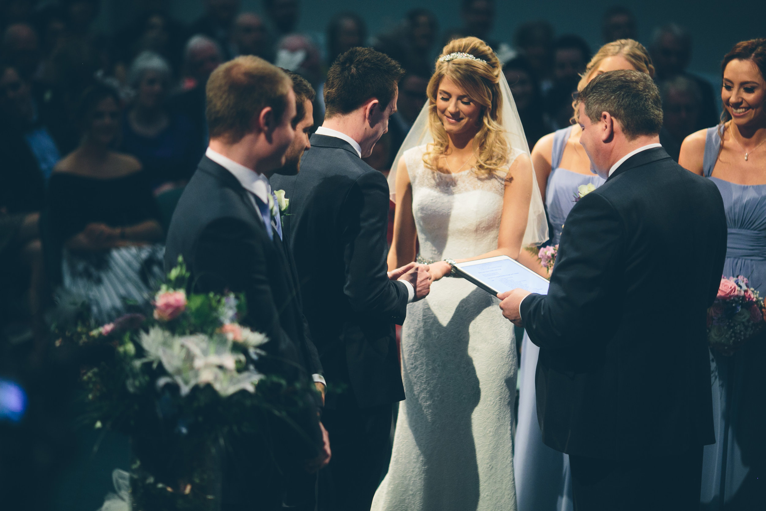 Bride and groom exchanging vows during a wedding ceremony, surrounded by groomsmen and bridesmaids, with an audience in the background.