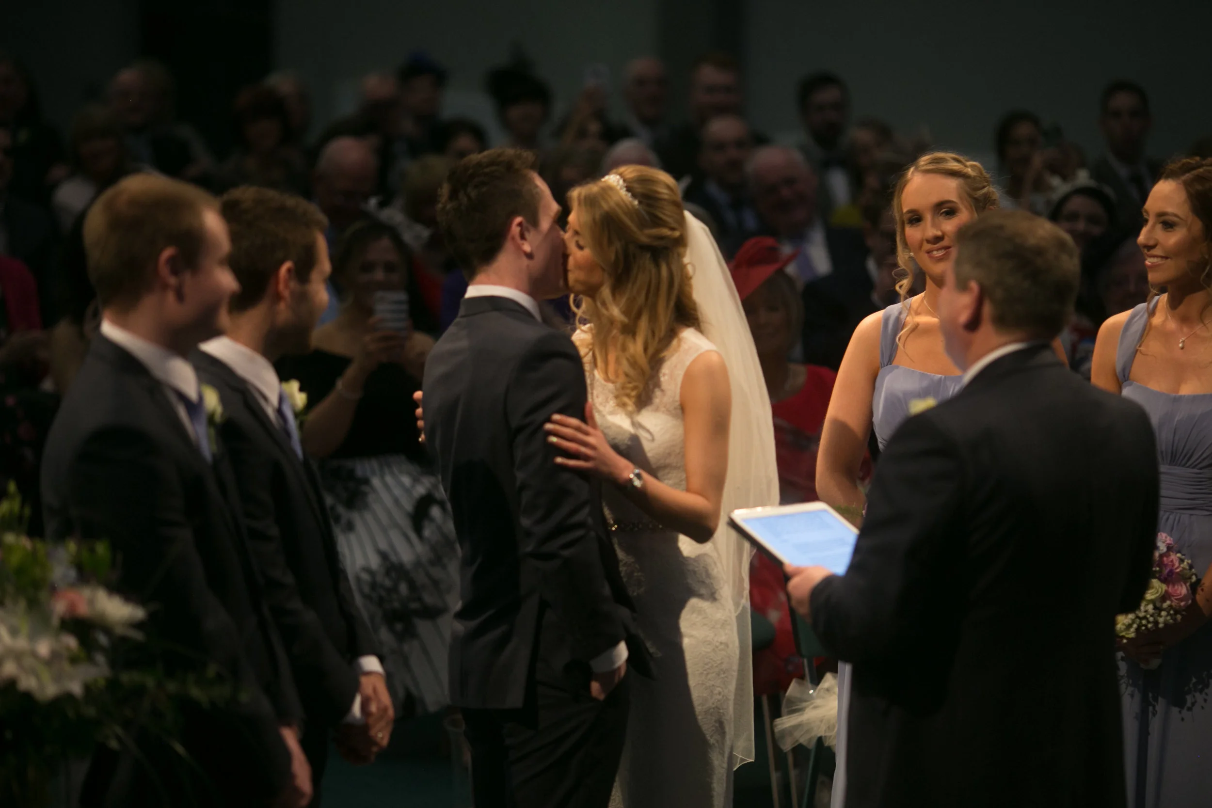Bride and groom kissing during wedding ceremony while officiant holds a tablet, with bridesmaids and groomsmen and guests watching in the background.
