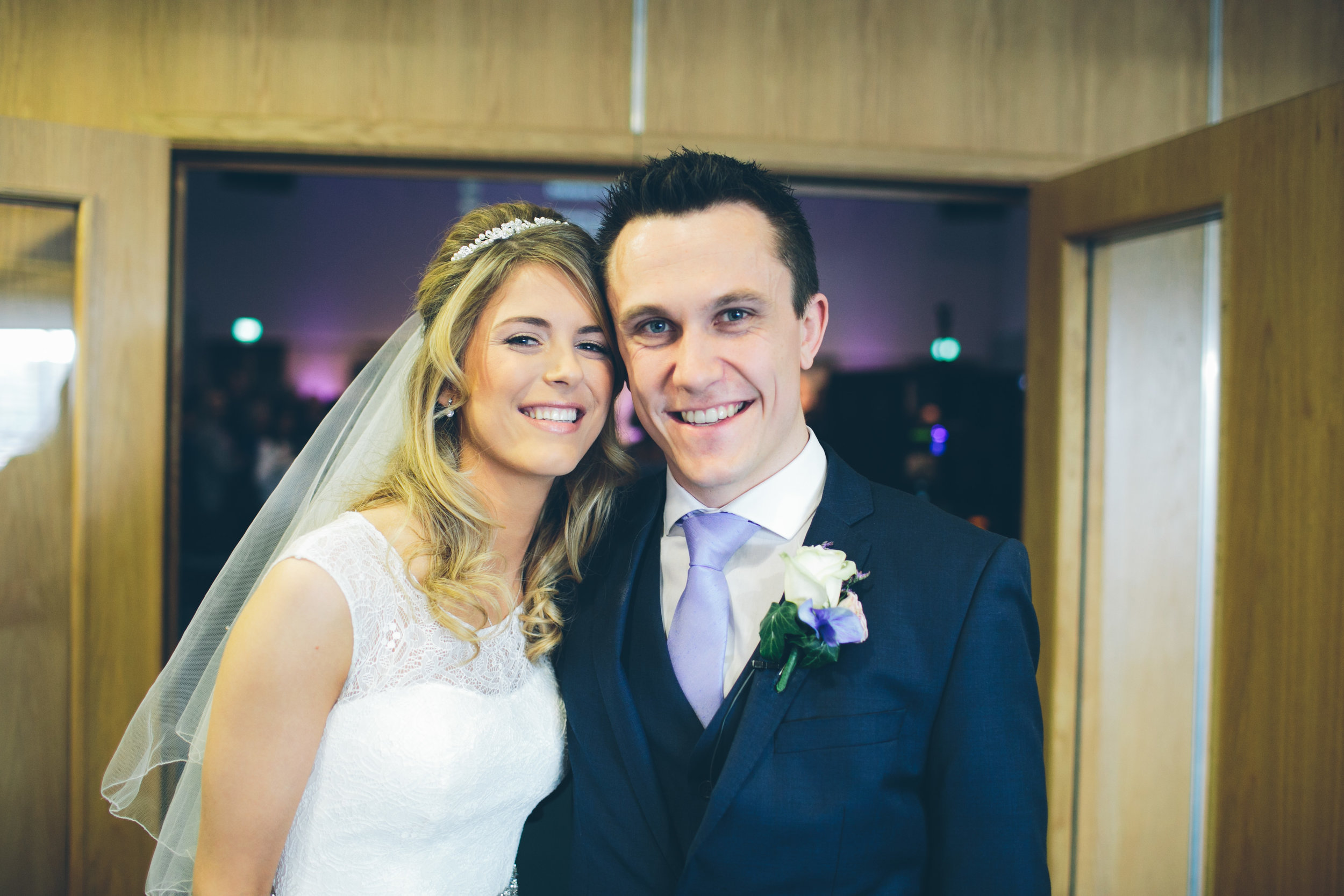 Bride and groom standing close together, smiling at the camera inside a wooden-paneled room.