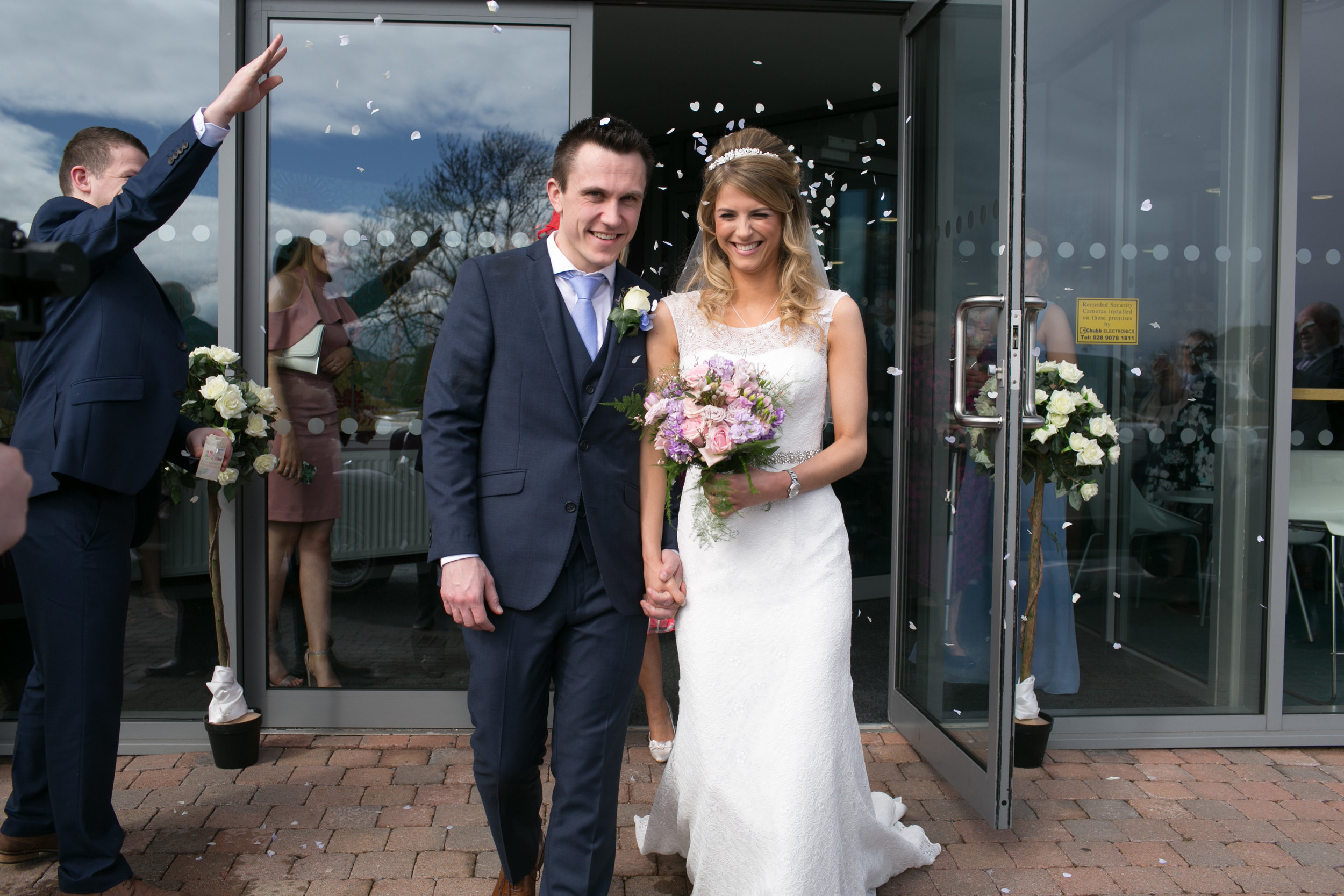 Bride and groom holding hands and smiling as they exit a building after their wedding, surrounded by flower decorations and confetti.