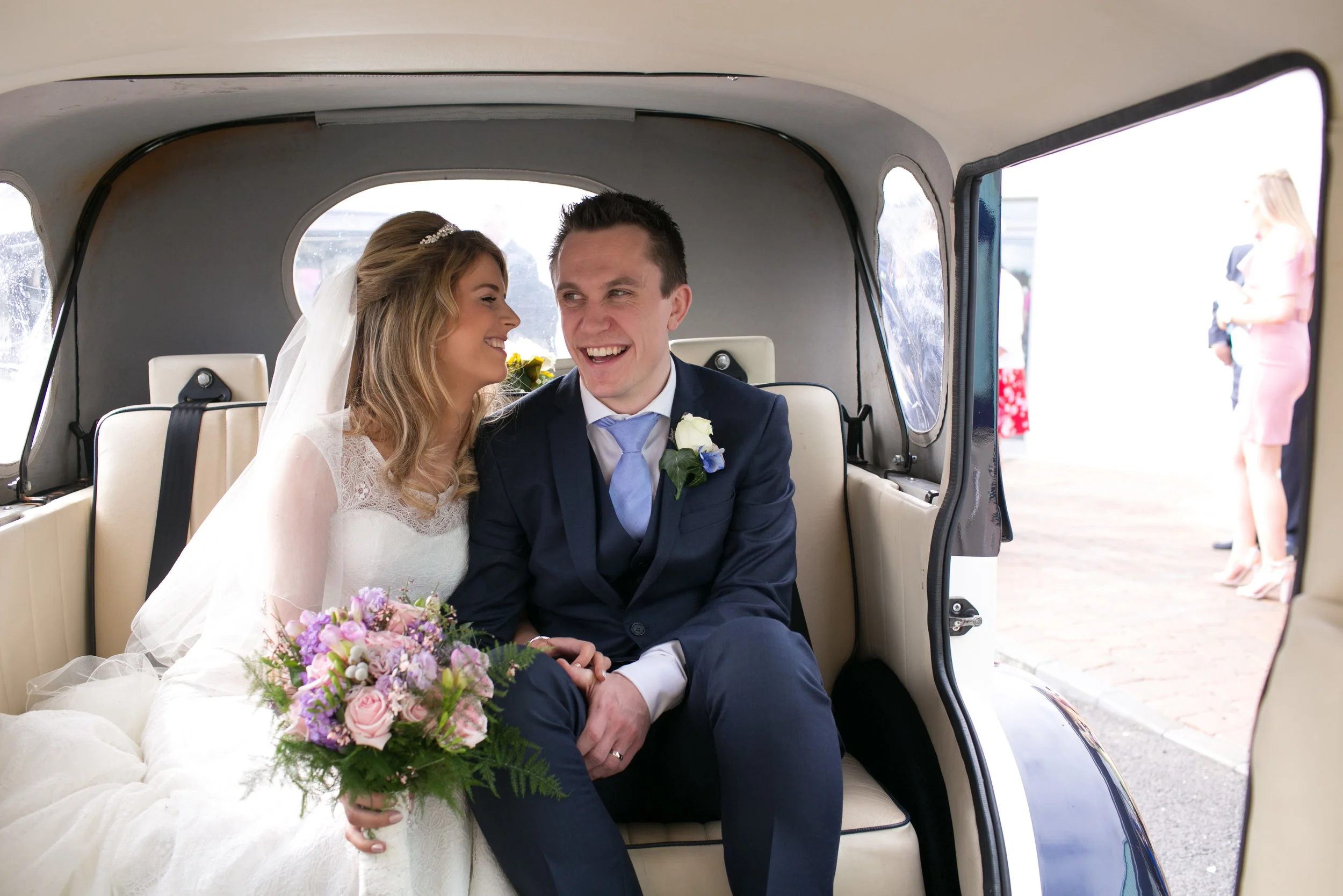 A bride and groom sitting inside a vintage vehicle, smiling and looking at each other. The bride holds a bouquet of pink and purple flowers, and the groom is wearing a dark suit with a light blue tie and boutonniere.
