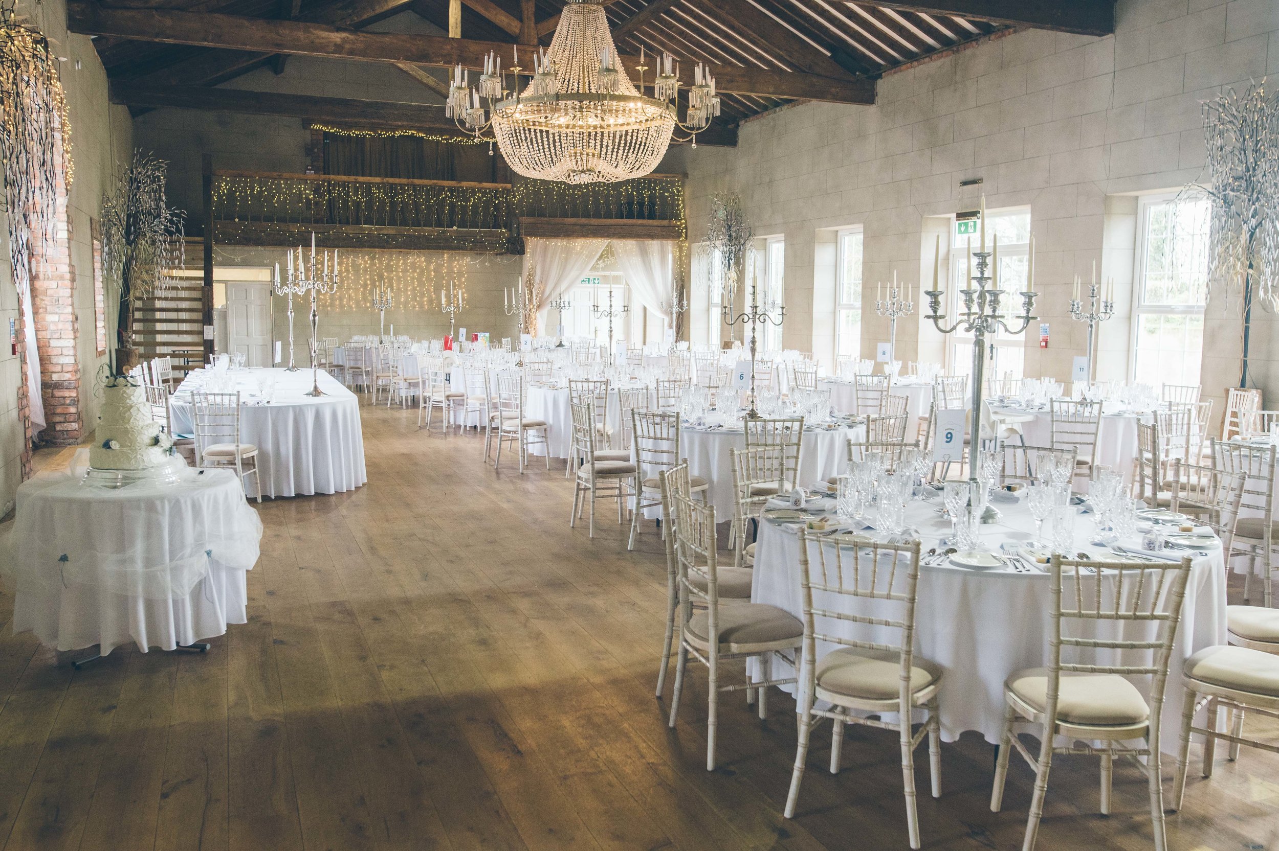 Elegant event hall decorated with white tablecloths, ornate candelabras, and string lights. Chandeliers hang from a rustic ceiling, with tall windows on the sides allowing natural light. A multi-tiered wedding cake is visible on a side table.