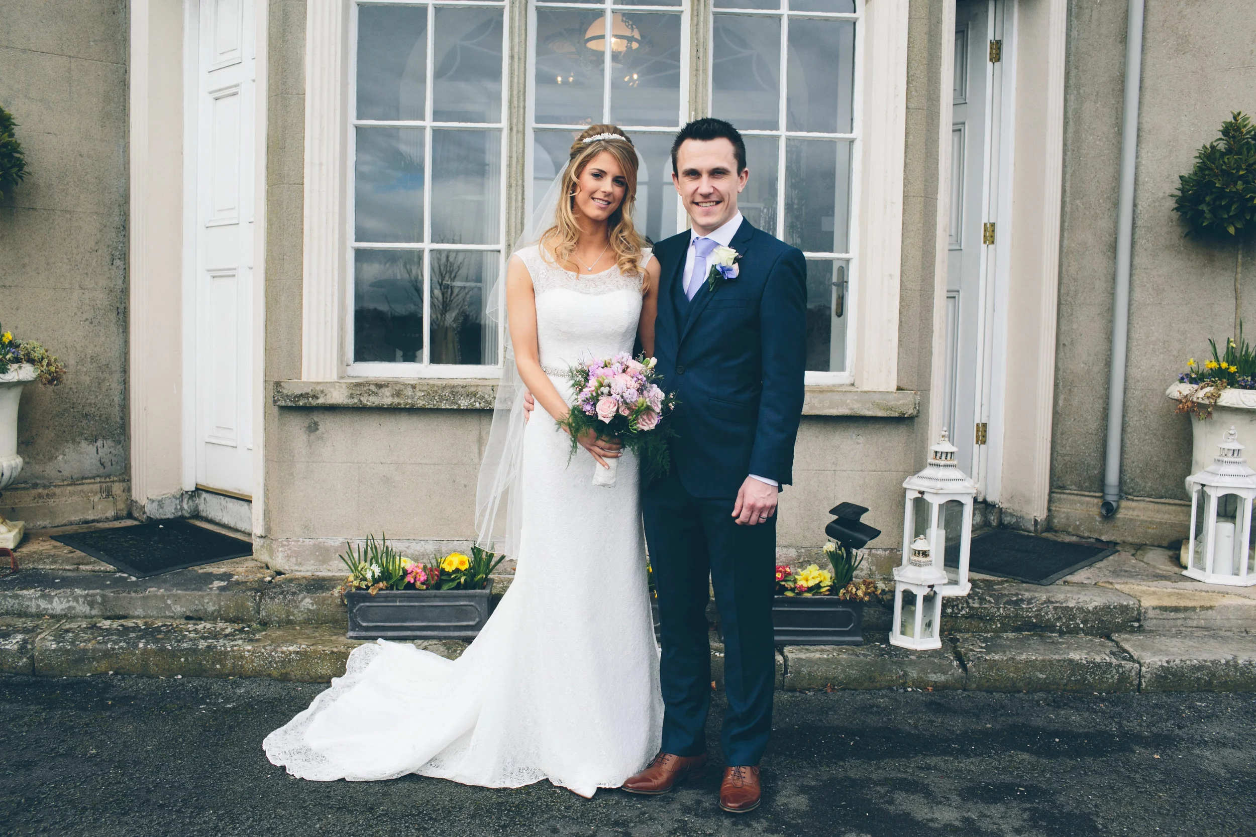 A bride in a white wedding dress and veil, holding a bouquet of pink and purple flowers, standing beside a groom in a dark blue suit, both smiling in front of a building with large windows and flower pots.