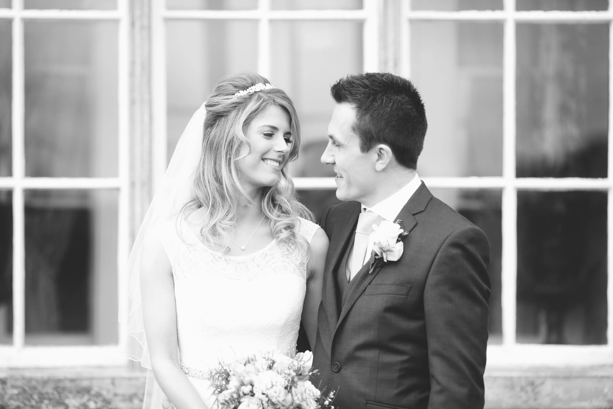 A black-and-white photo of a bride and groom facing each other, smiling, with a large window behind them.