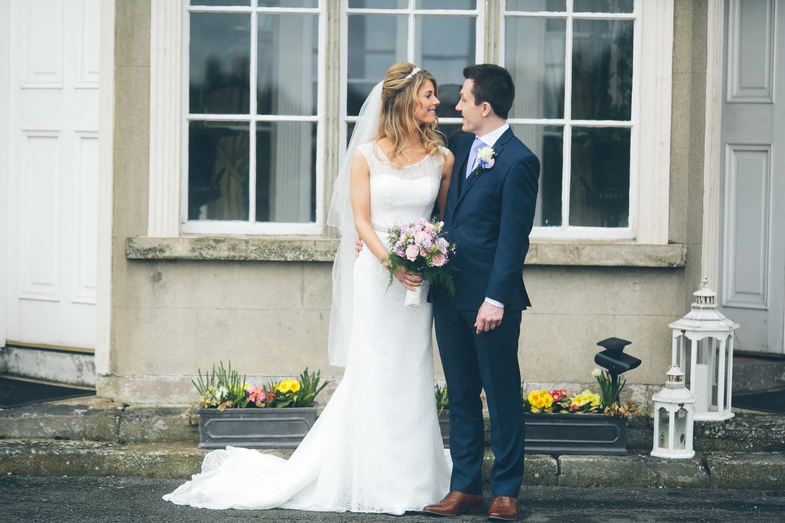 A bride and groom stand close together outside a building with large windows, looking into each other's eyes. The bride is holding a bouquet of pink and purple flowers, wearing a white wedding dress and a veil. The groom is dressed in a dark blue sui