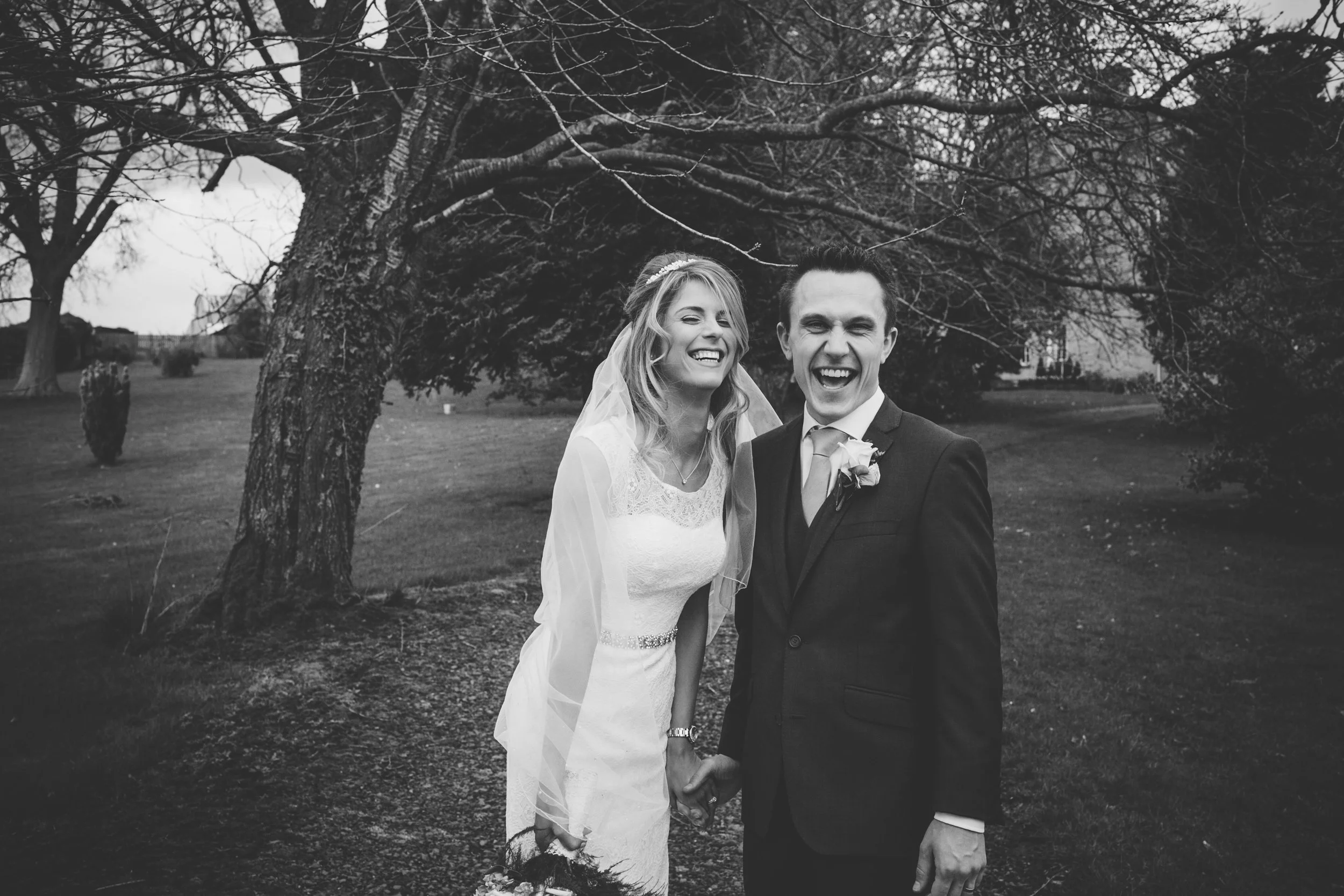 A black and white photo of a smiling bride and groom holding hands outdoors under a large, leafless tree, on a cloudy day.