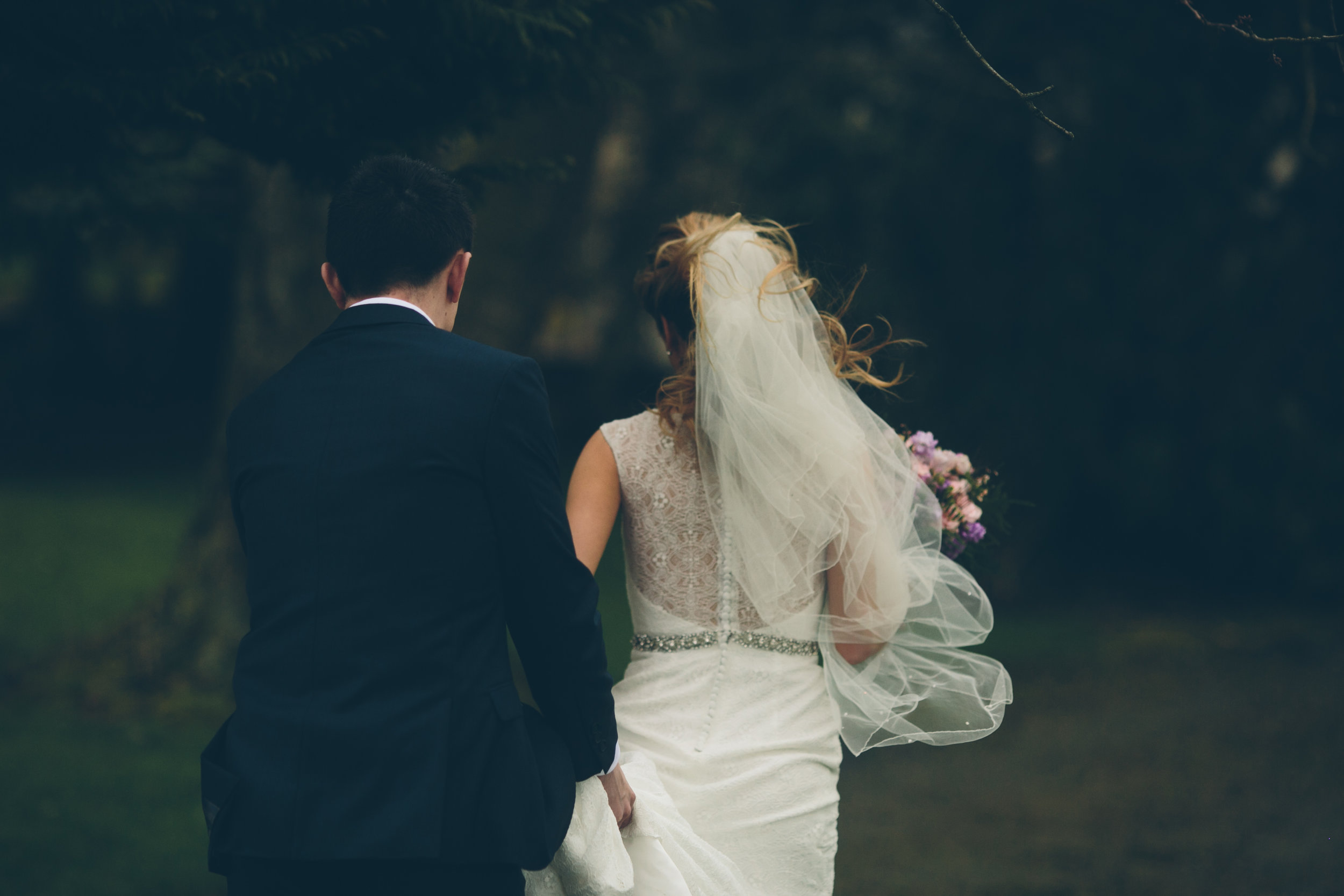A bride and groom walking outdoors, seen from the back, with the bride holding a bouquet, surrounded by trees.