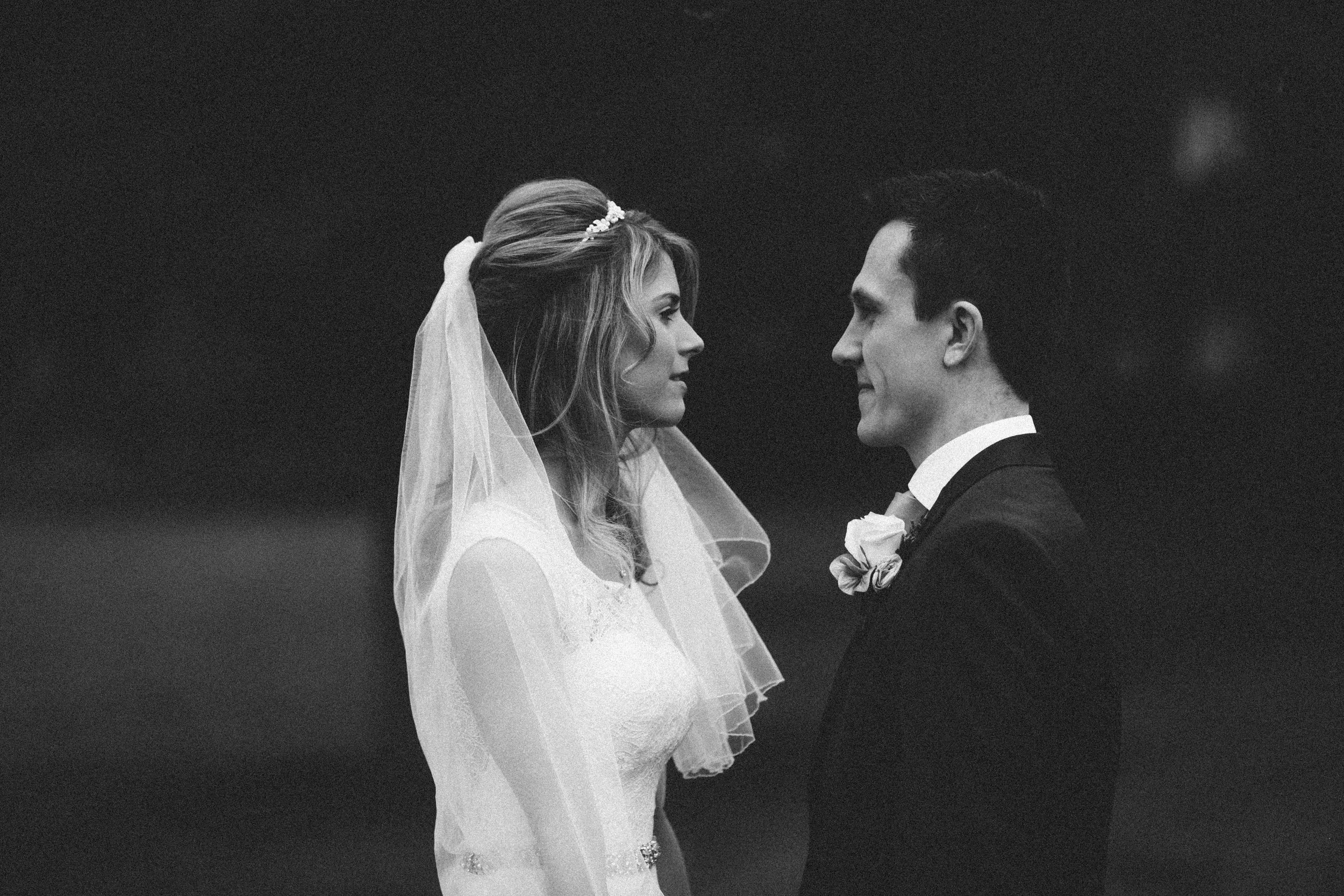 Black and white photo of a bride and groom facing each other, wedding attire, bride with veil, groom with boutonniere, intimate moment during wedding ceremony.