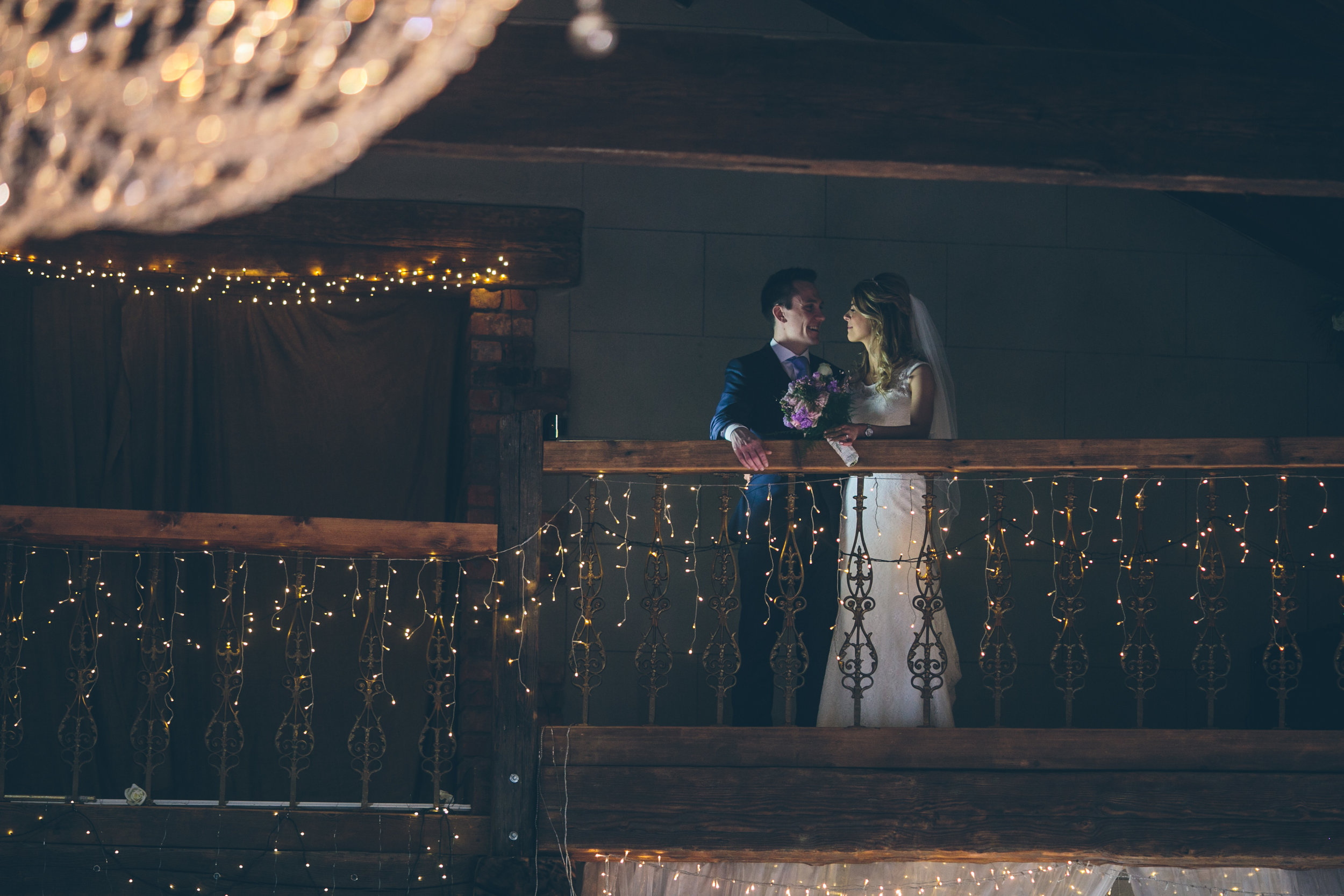 Bride and groom standing on a decorated balcony, smiling at each other, with string lights hanging around them.