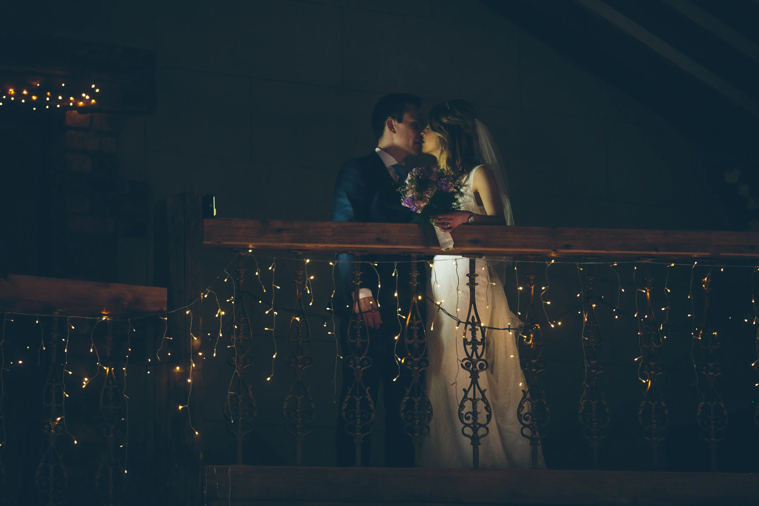 A bride and groom sharing a kiss on a balcony decorated with string lights during a wedding celebration.