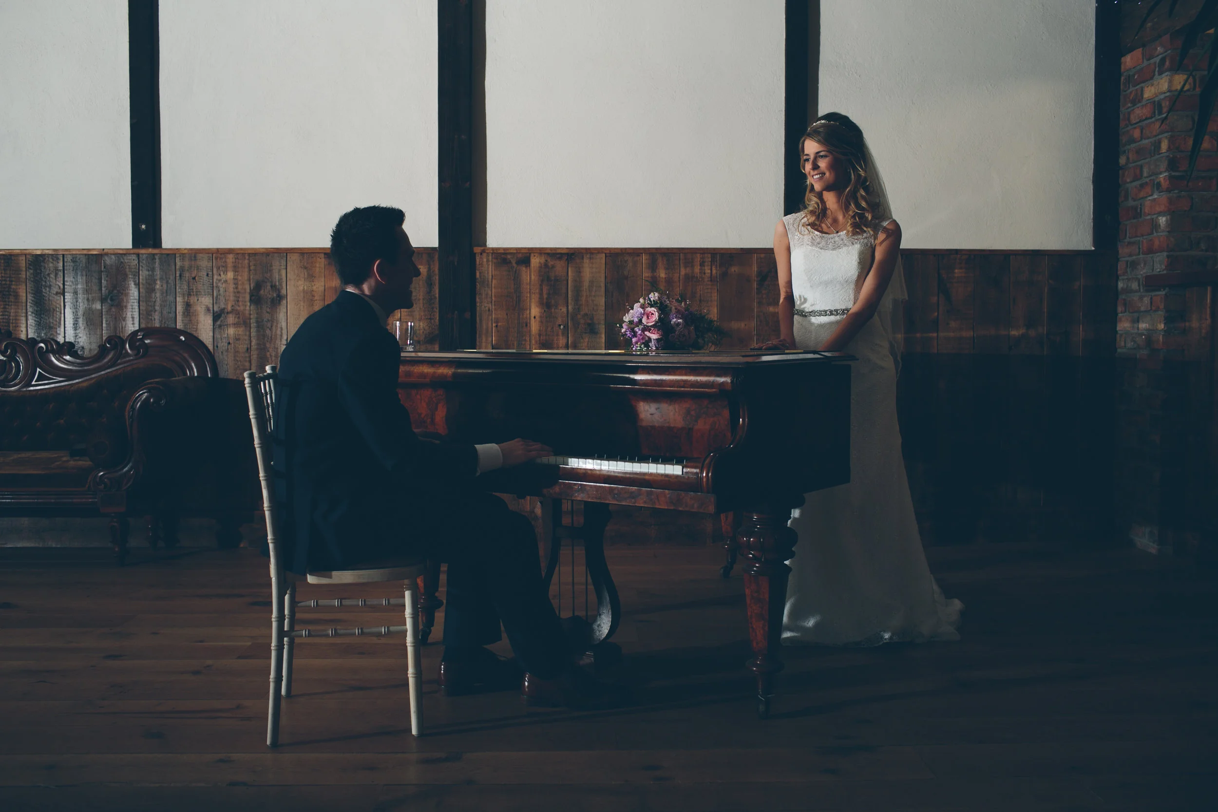 A bride in a white wedding dress stands near a pianist playing a grand piano in a rustic indoor setting.