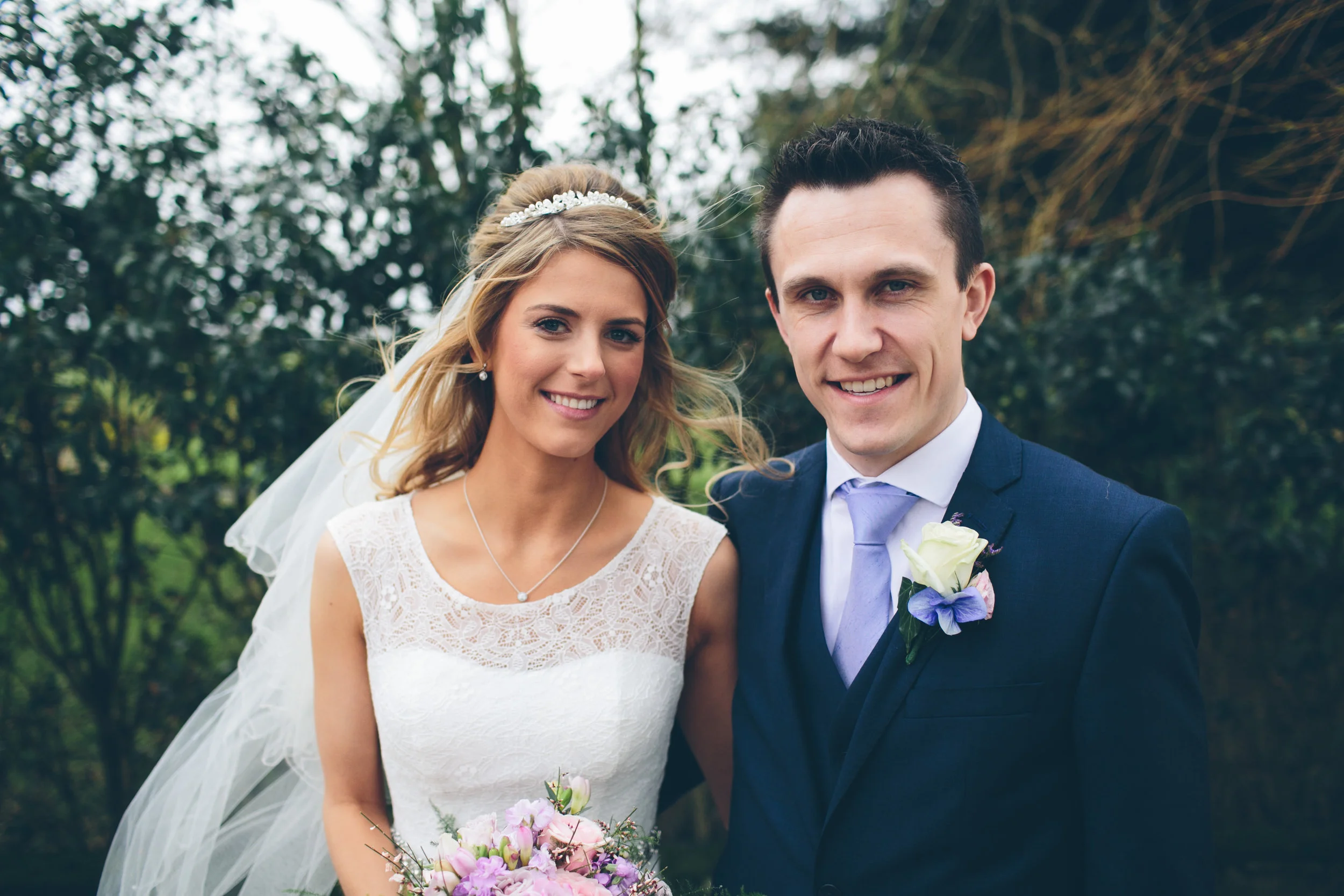 A bride and groom smiling together outdoors on their wedding day, with trees in the background. The bride wears a lace wedding dress and veil, holding a bouquet of pink and purple flowers. The groom is dressed in a dark blue suit with a lavender tie 