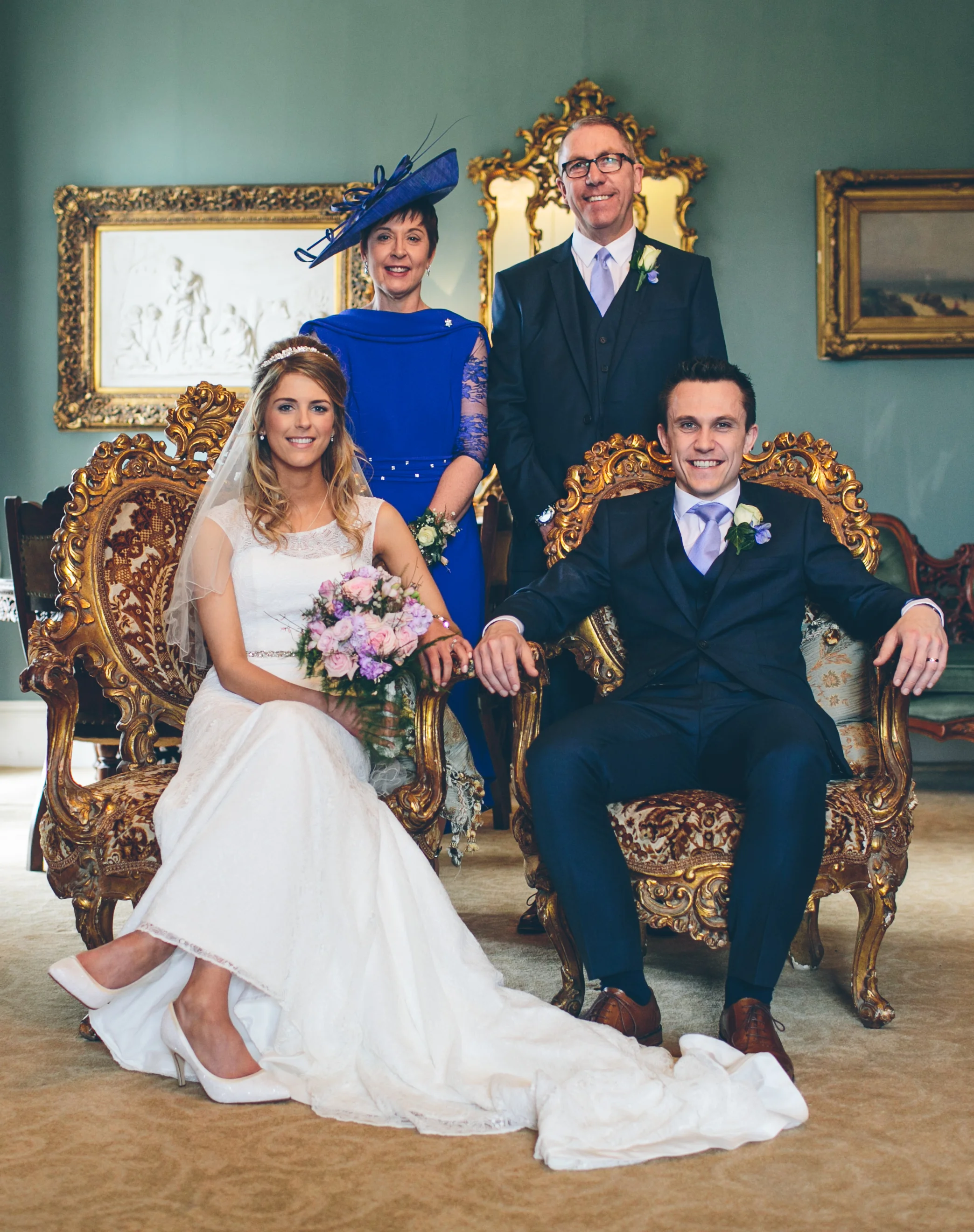 A wedding photo featuring a bride and groom seated on an ornate vintage sofa, with two adults standing behind them in a formal setting with classic paintings and gold-framed mirrors.
