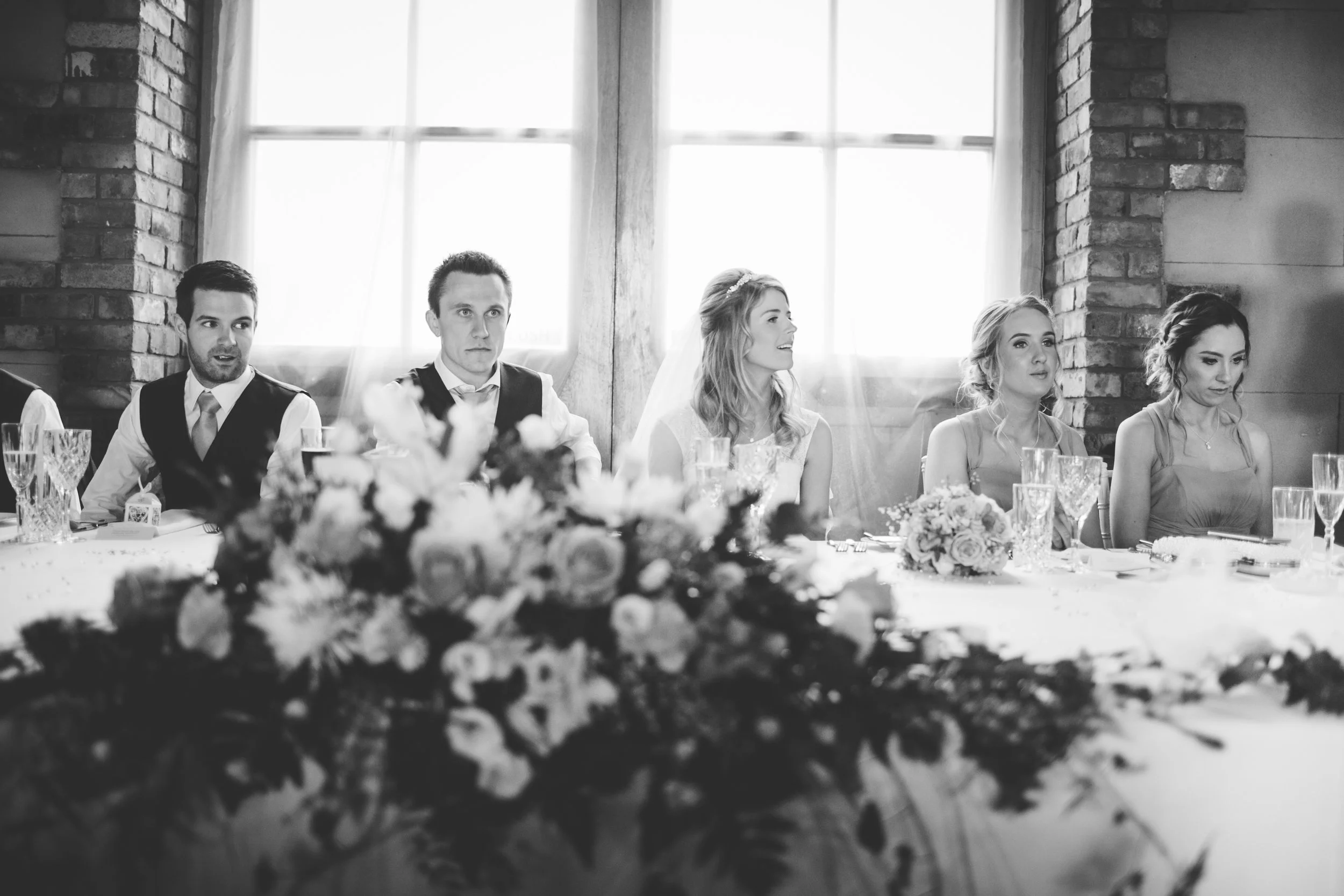 Black and white photo of five people sitting at a long table during a wedding reception. A woman wearing a wedding dress with a veil is in the center, laughing and looking to the right. To her left, a woman in a sleeveless dress looks straight ahead;