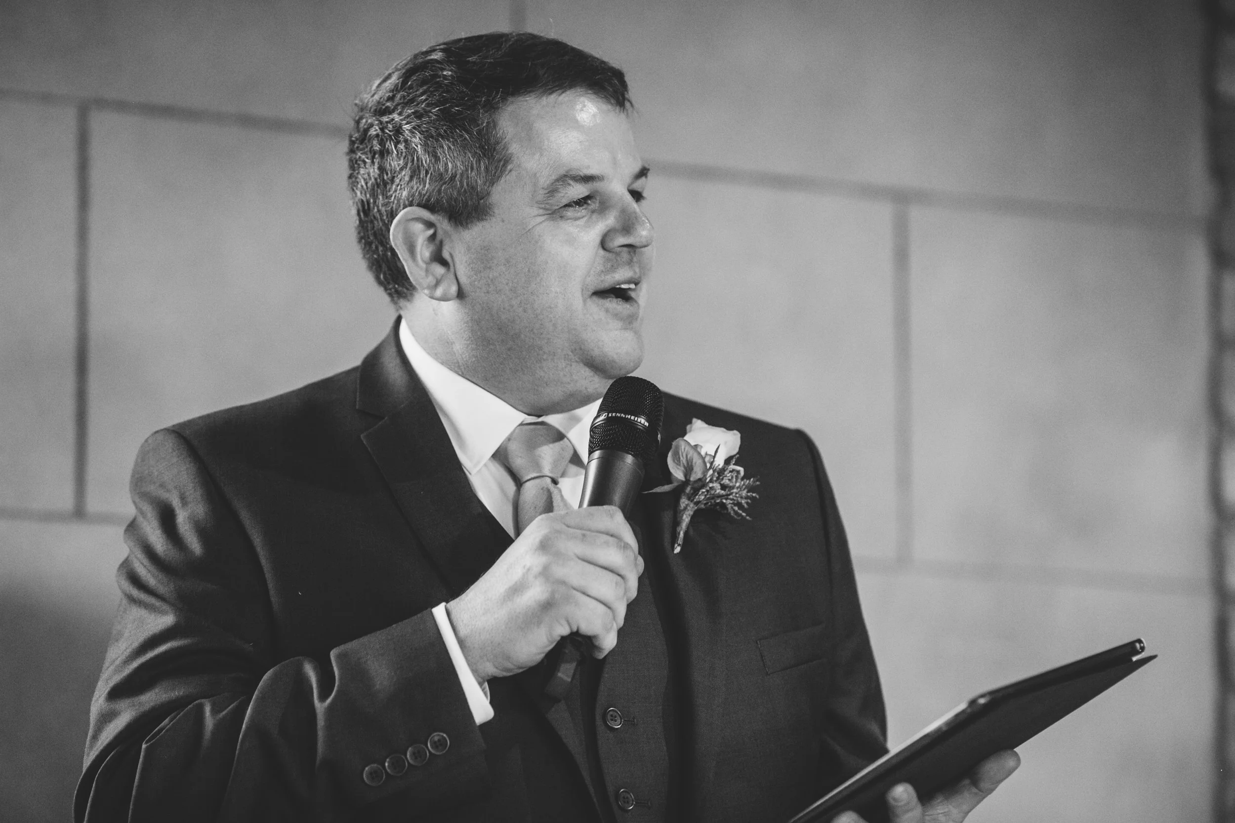 A man in a suit holding a microphone and a tablet, speaking at an event.