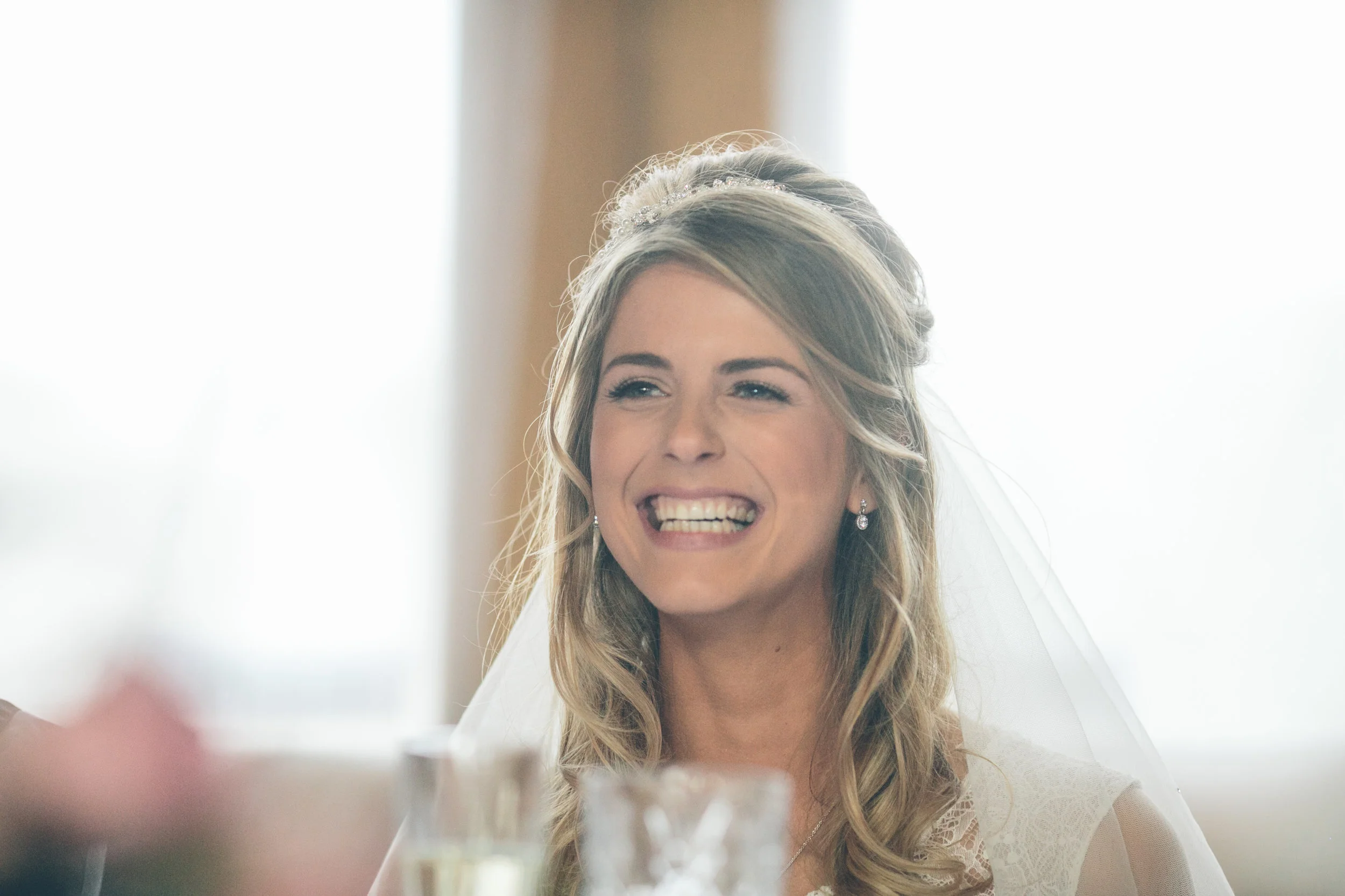 A bride smiling happily with her eyes slightly squinted, wearing a wedding dress and earrings, with a bridal veil, indoors with natural light.