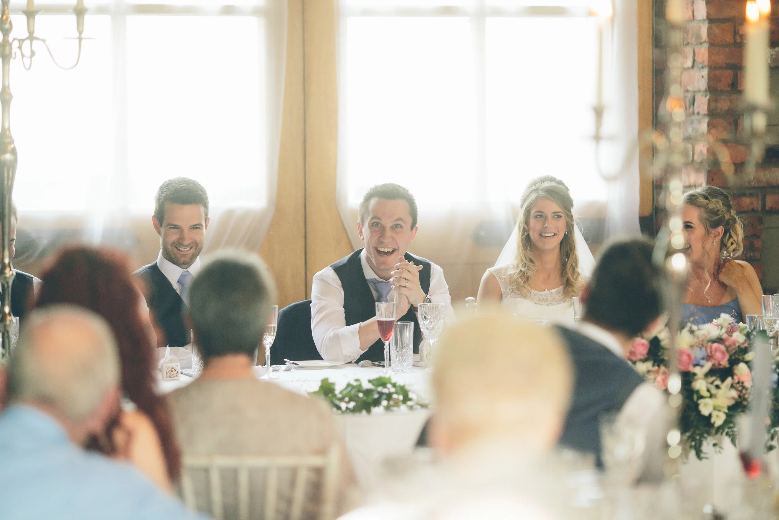 Group of wedding guests sitting at a banquet table, watching and smiling as the bride and groom are seated at the head table during a wedding celebration.