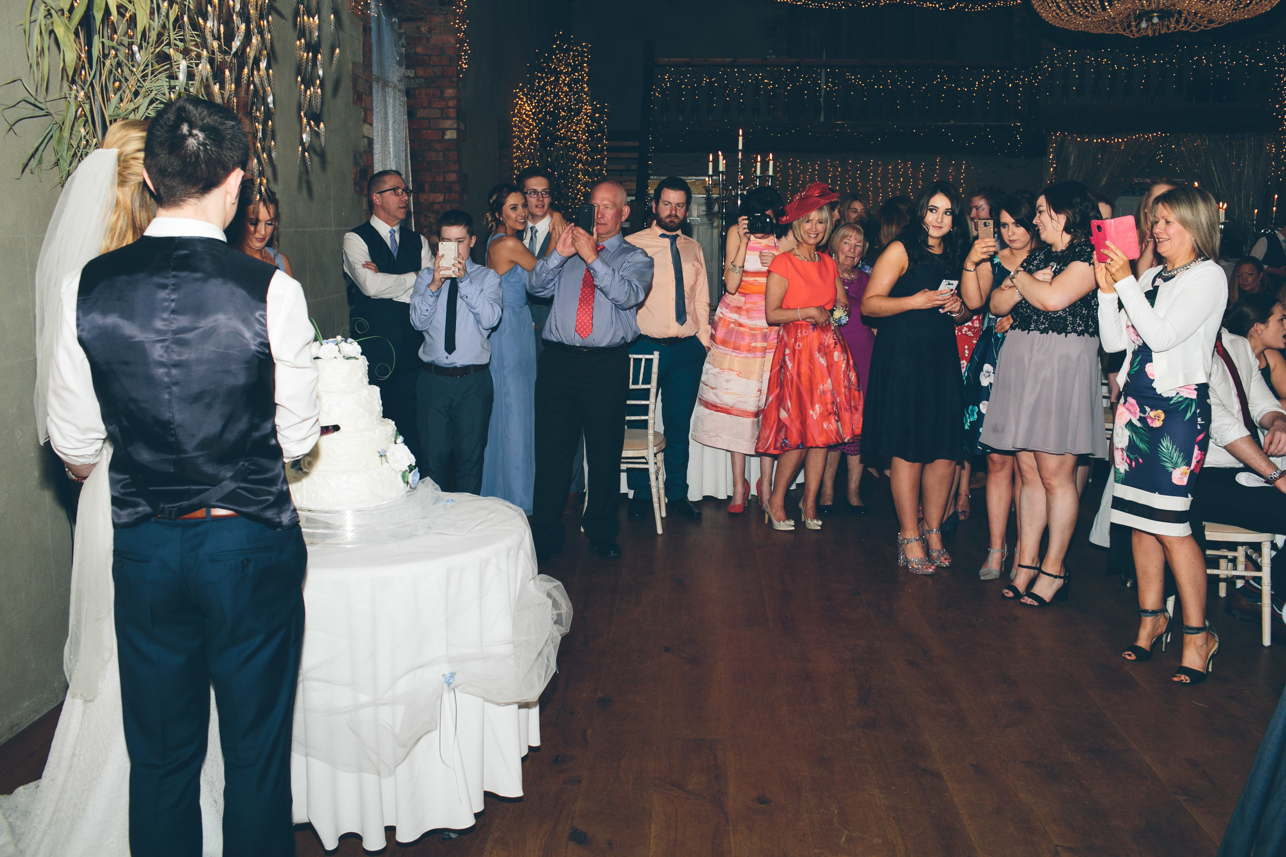 A wedding reception with a bride and groom standing near a multi-tiered wedding cake, surrounded by guests taking photos and videos. The scene is decorated with string lights and candles.