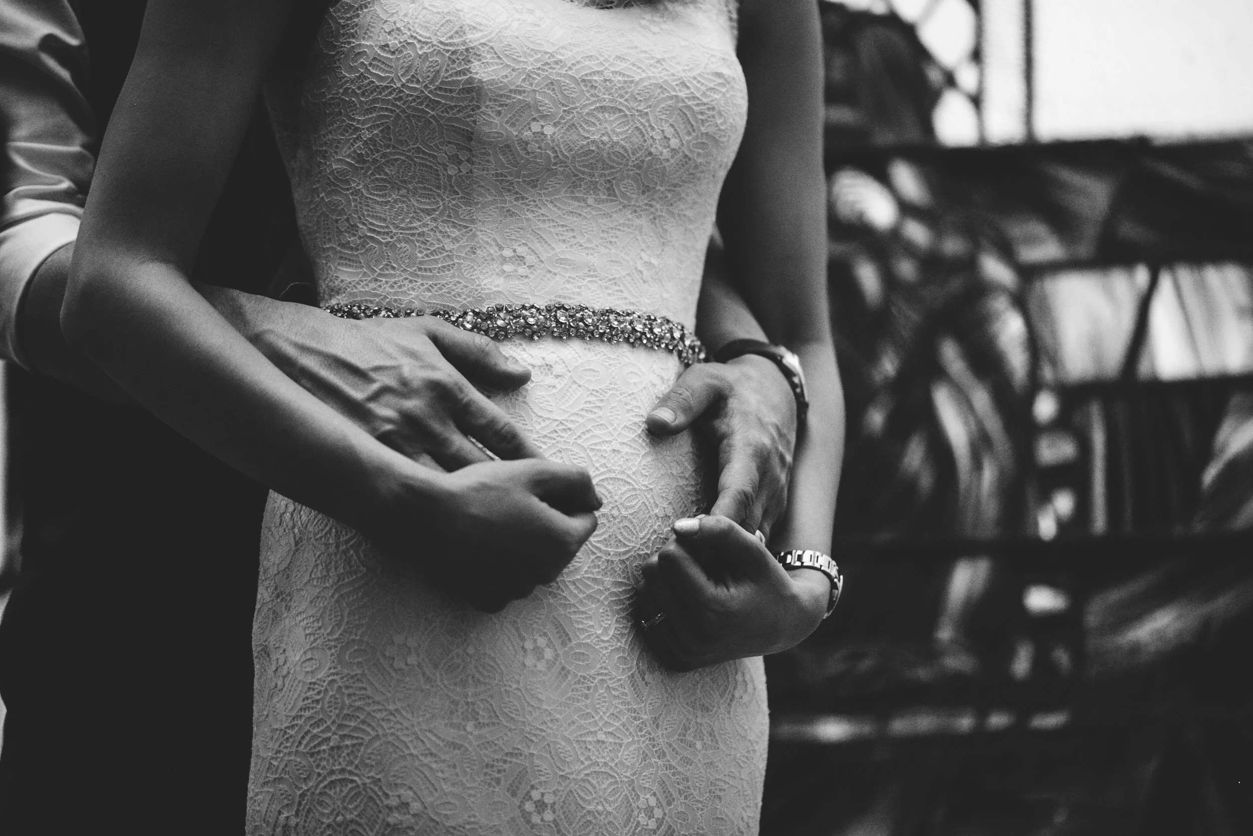 Close-up of a bride in a lace wedding dress with a bejeweled belt, holding her belly, supported by two hands, likely a partner's, with a jewelry watch and ring visible. The background is blurred, showing shelves with plants and books.