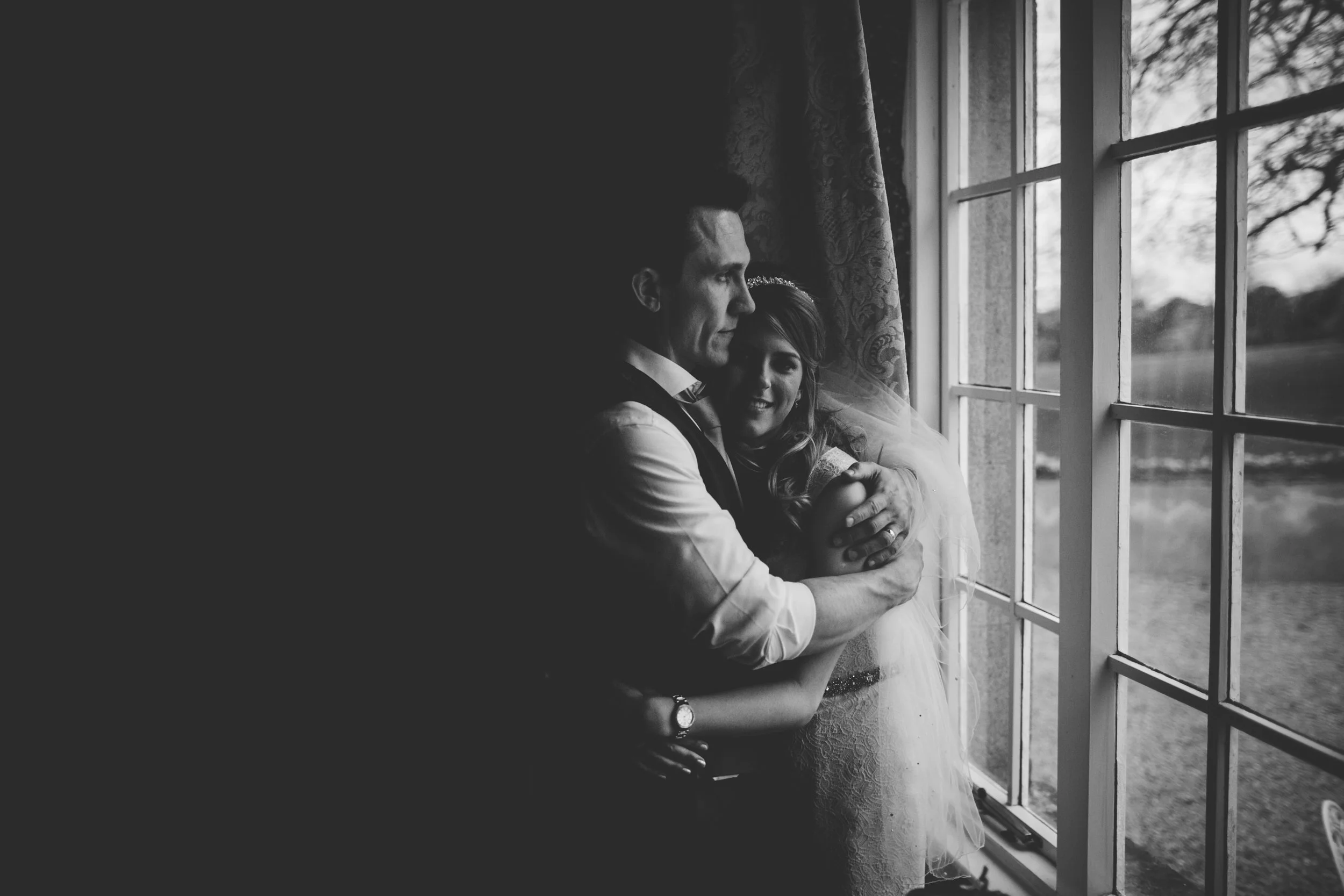 A black-and-white photo of a couple standing by a window, embracing each other. The man is looking out the window while the woman, smiling, leans into his shoulder. The window reveals a scenic outdoor view.