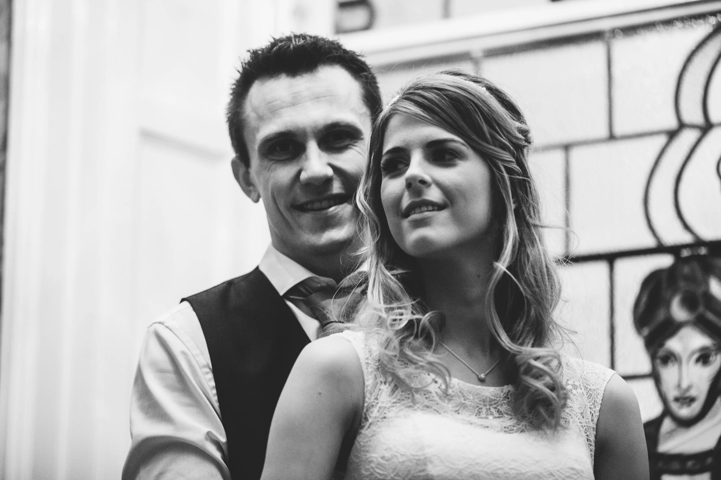 A black-and-white photo of a smiling man in a vest and tie standing closely behind a young woman with wavy hair and a lace dress, with a city mural featuring a woman's face in the background.