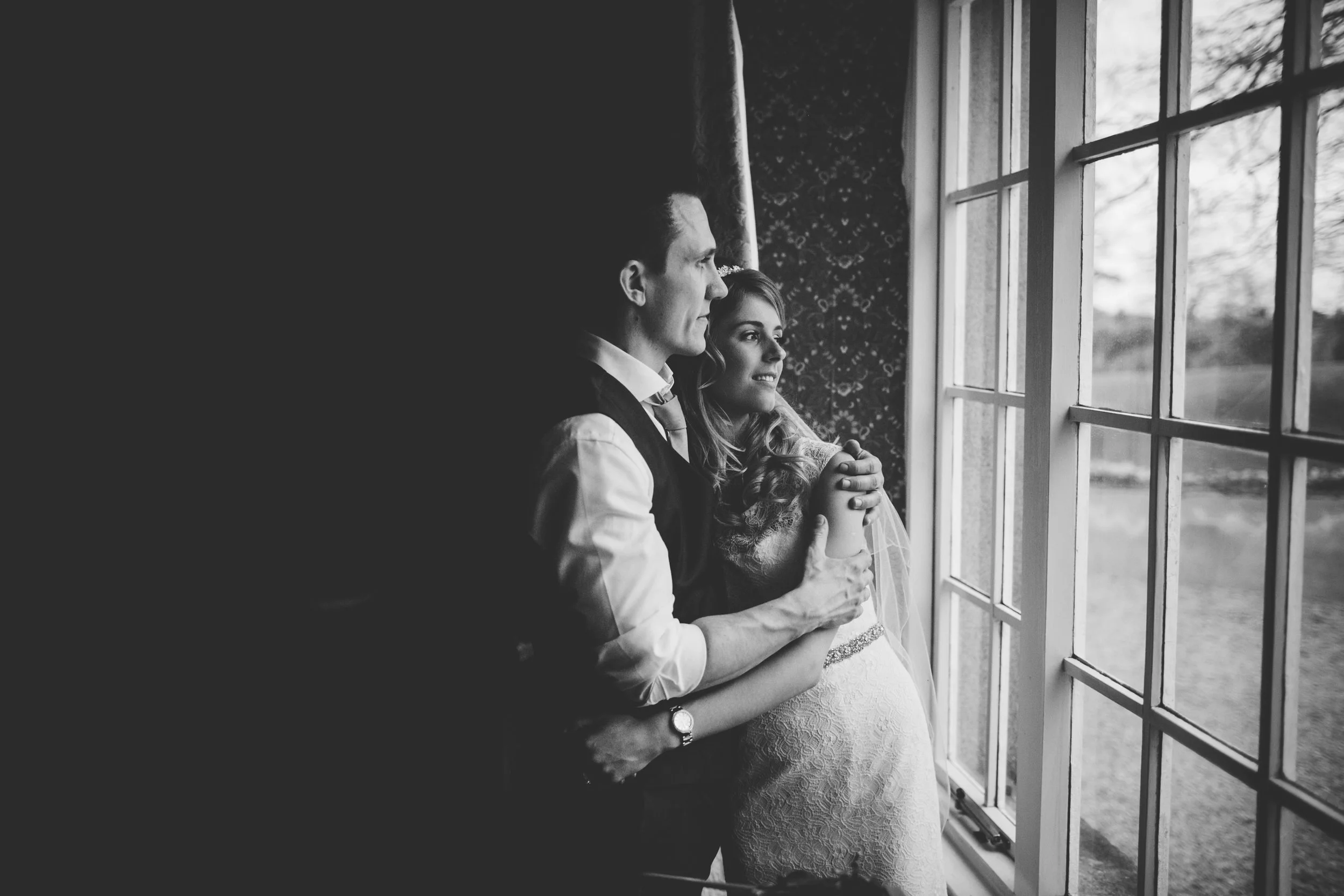 A couple, dressed in wedding attire, standing by a window and looking outside, with the woman holding a doll, in black and white photo.