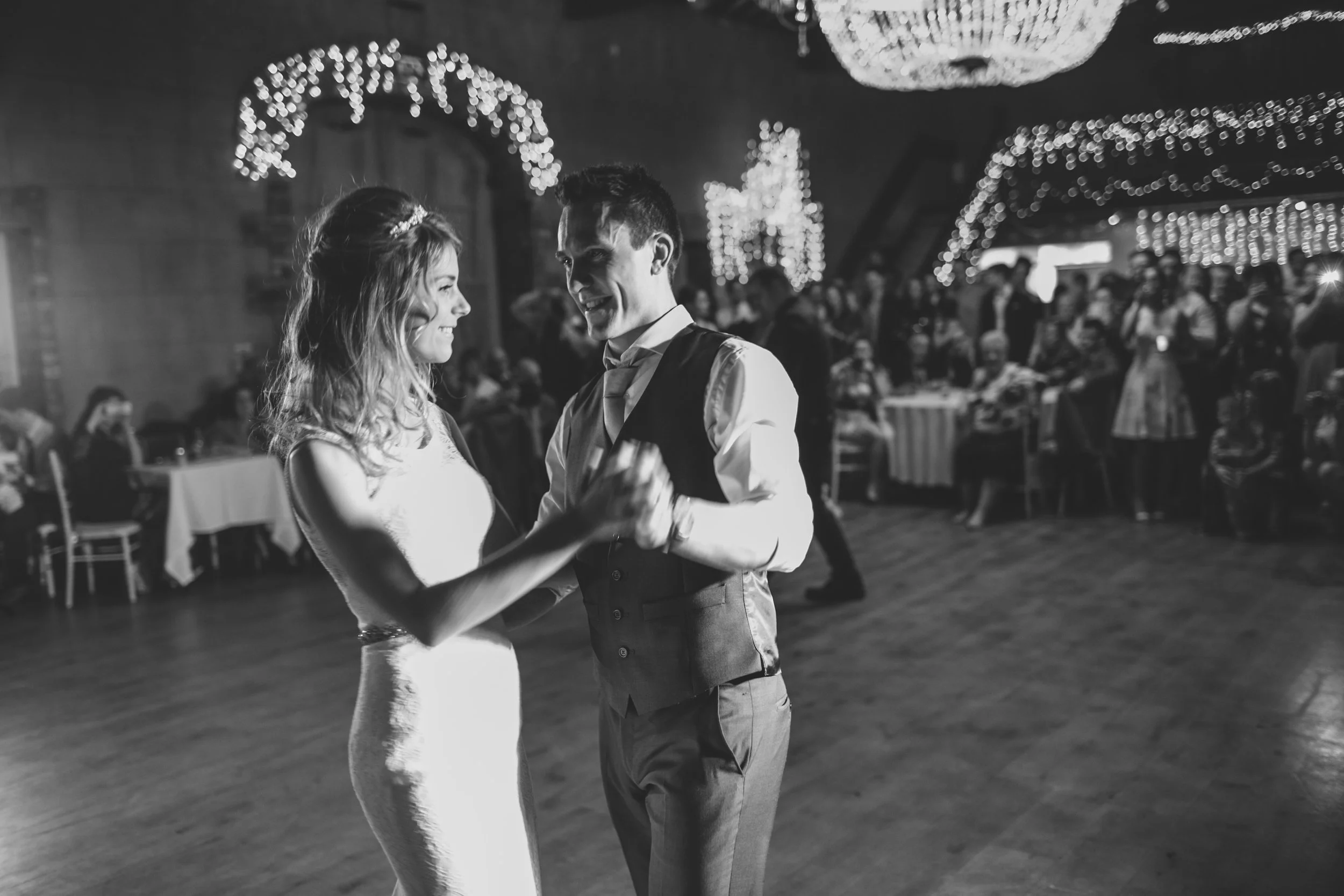 A black-and-white photo of a bride and groom dancing at their wedding reception, with guests seated at tables in the background and string lights decorating the venue.