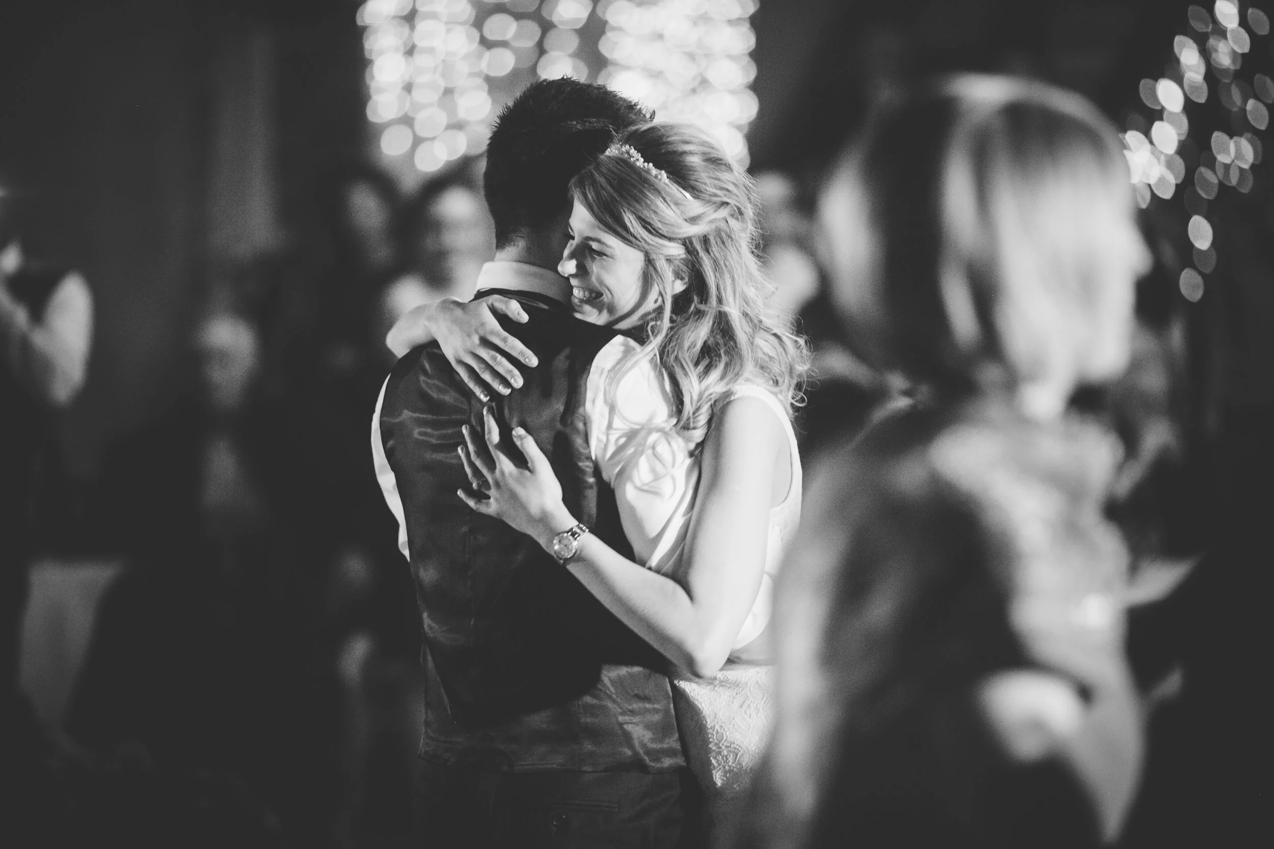 A black and white photo of a couple dancing at their wedding, with the woman smiling and embracing the man, surrounded by blurred guests and string lights in the background.