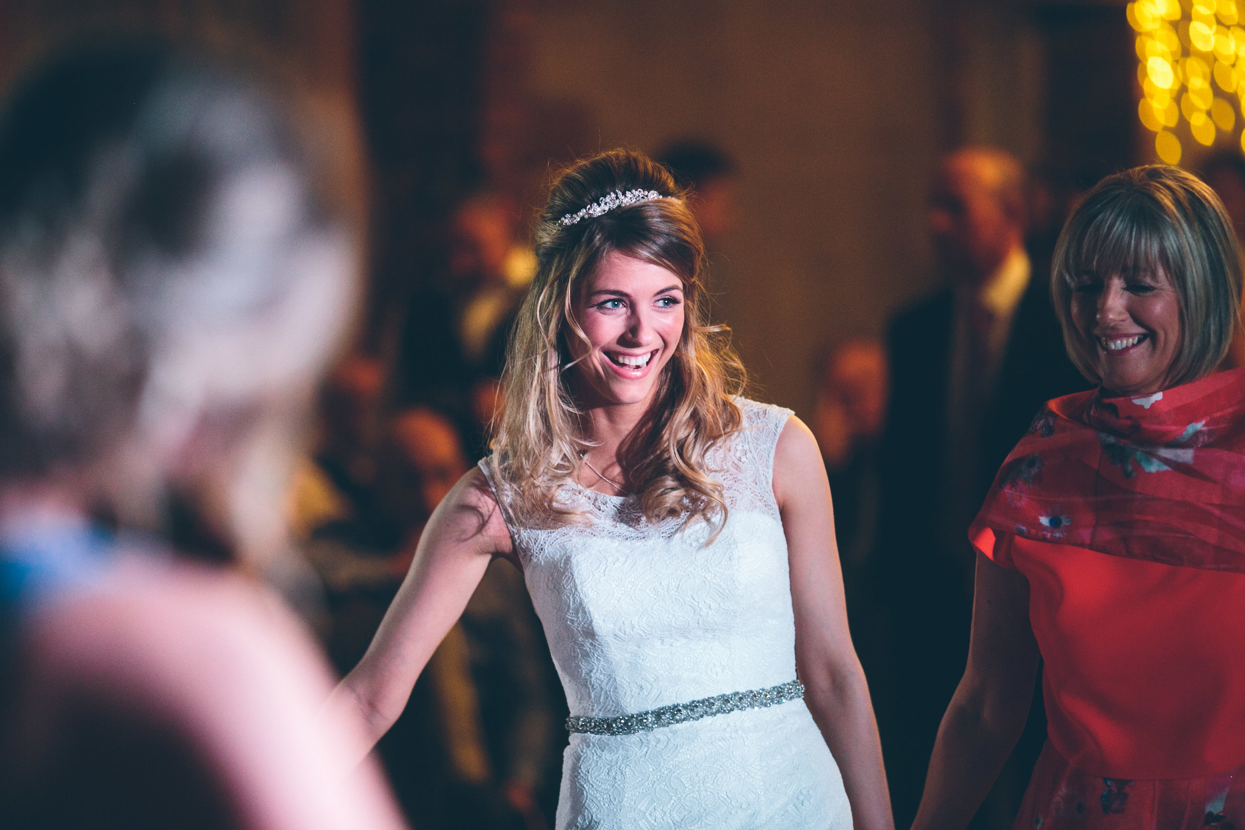 A woman in a white wedding dress and tiara smiling and dancing with others at a wedding reception.