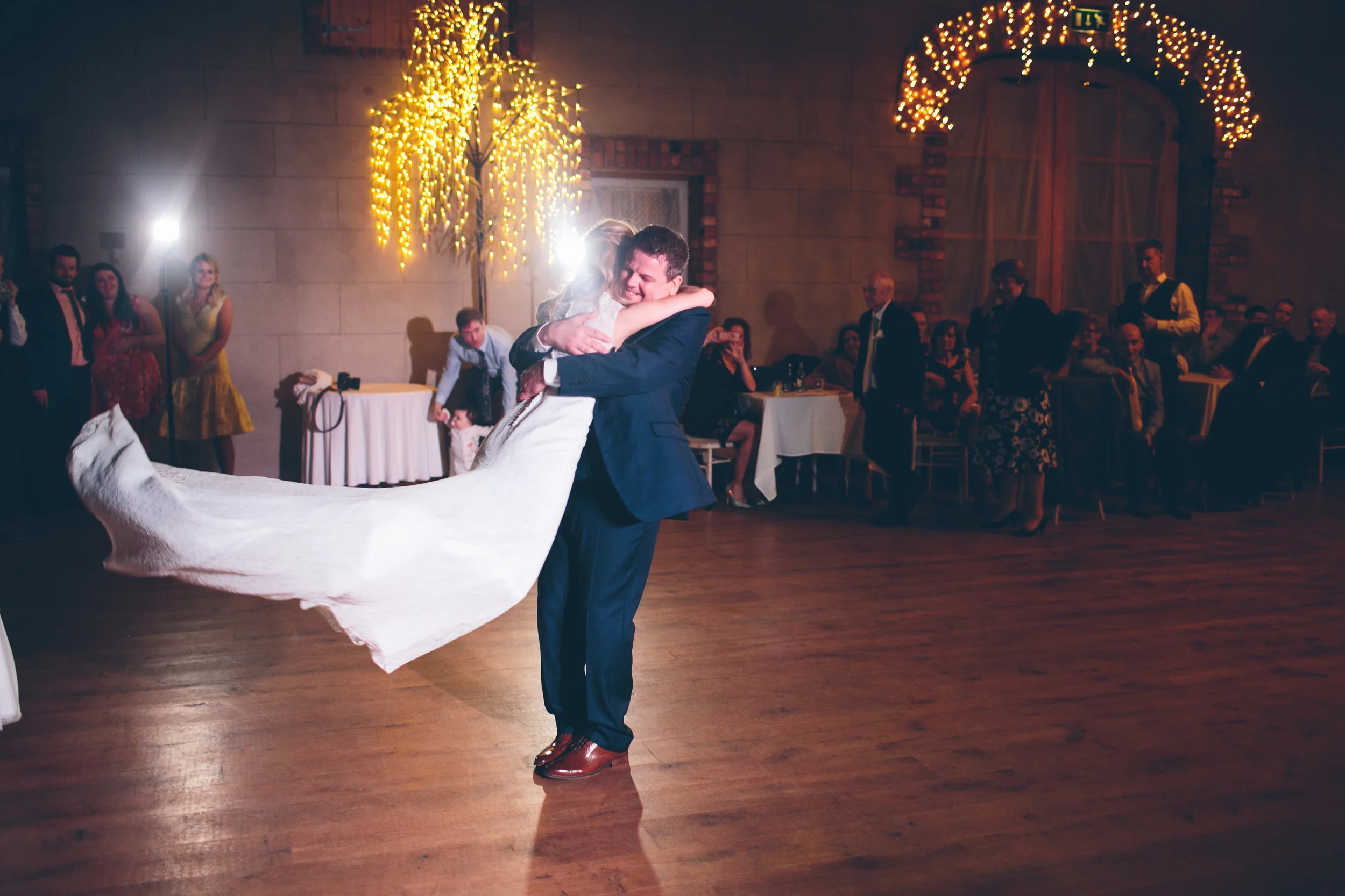 A bride and groom dancing closely at their wedding reception, with the groom dipping the bride. Guests are watching and smiling in the background, decorated with warm string lights and a rustic indoor setting.