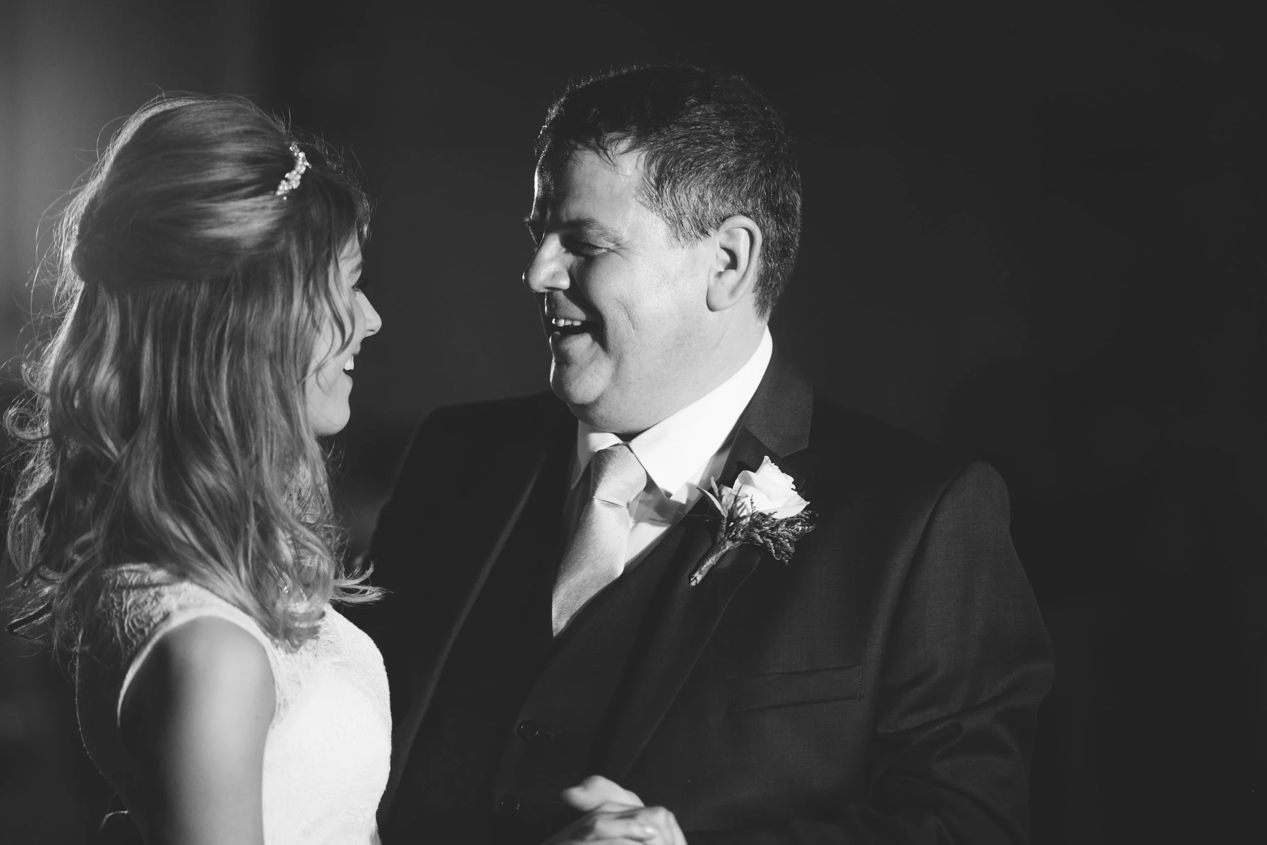 A black and white photo of a groom and bride dancing closely, facing each other, smiling at each other, with the groom wearing a suit with a boutonniere and the bride in a sleeveless dress with wavy hair.