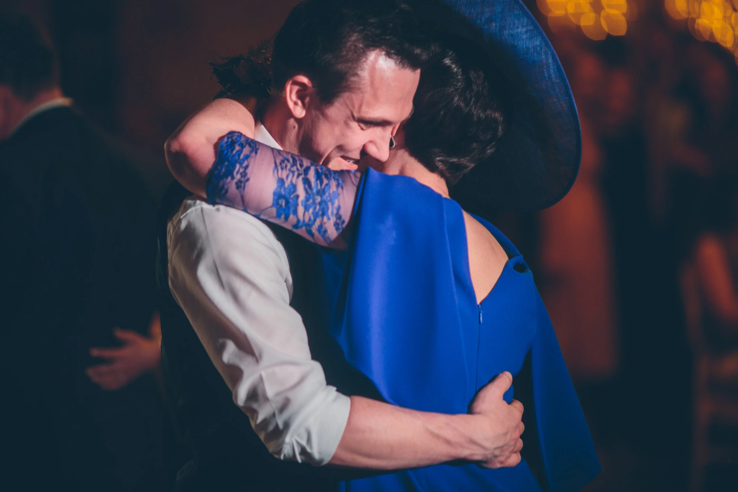 A man and woman hugging warmly at a social event, with the woman wearing a blue dress and a wide-brimmed hat, in a dimly lit setting with blurred background lights.