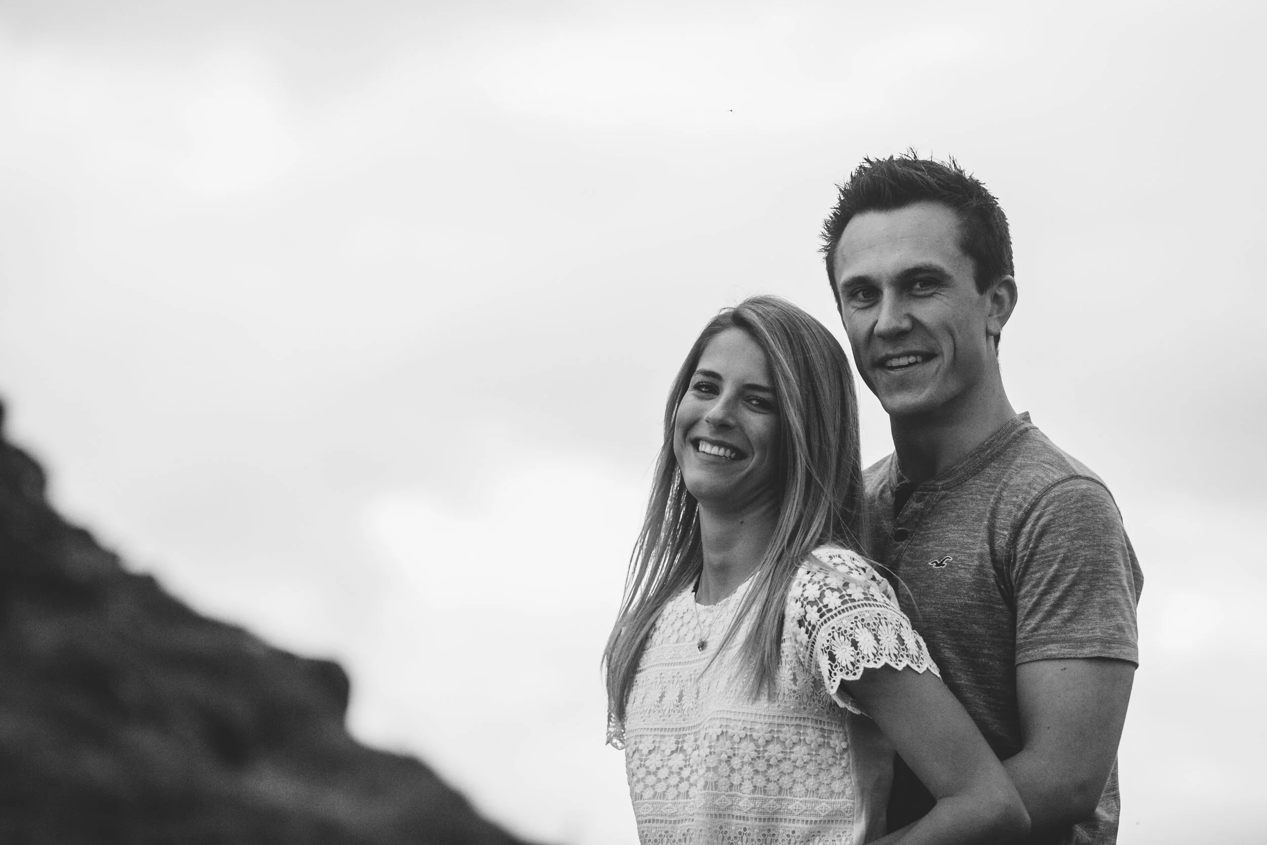 A black and white photo of a smiling young couple outdoors, with the woman having long hair and wearing a lace top, and the man having short hair and wearing a casual t-shirt, standing close together against a cloudy sky.