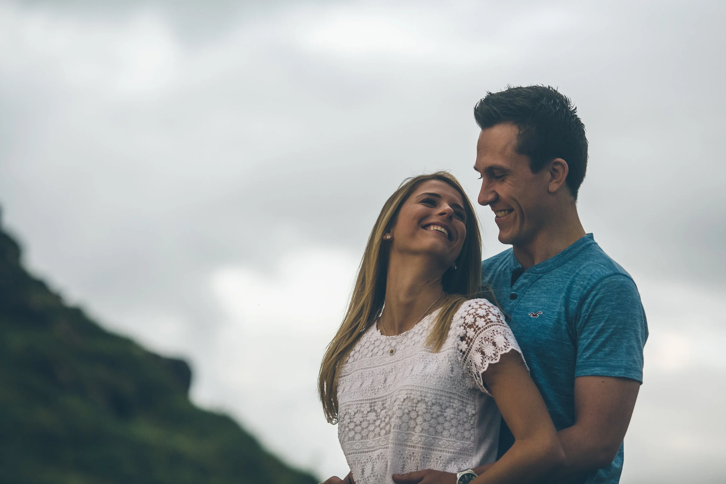 A smiling woman and man in casual clothes looking at each other outdoors on a cloudy day.