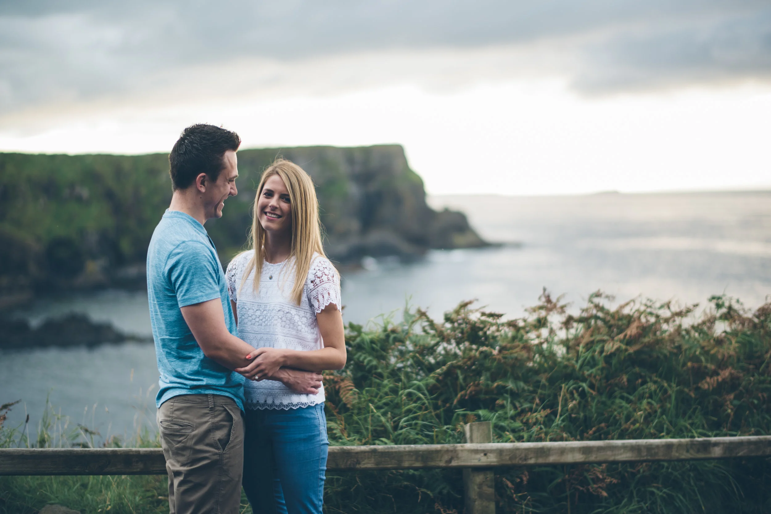 A young couple standing on a wooden fence by the coast, with cliffs and ocean in the background, smiling and holding hands.