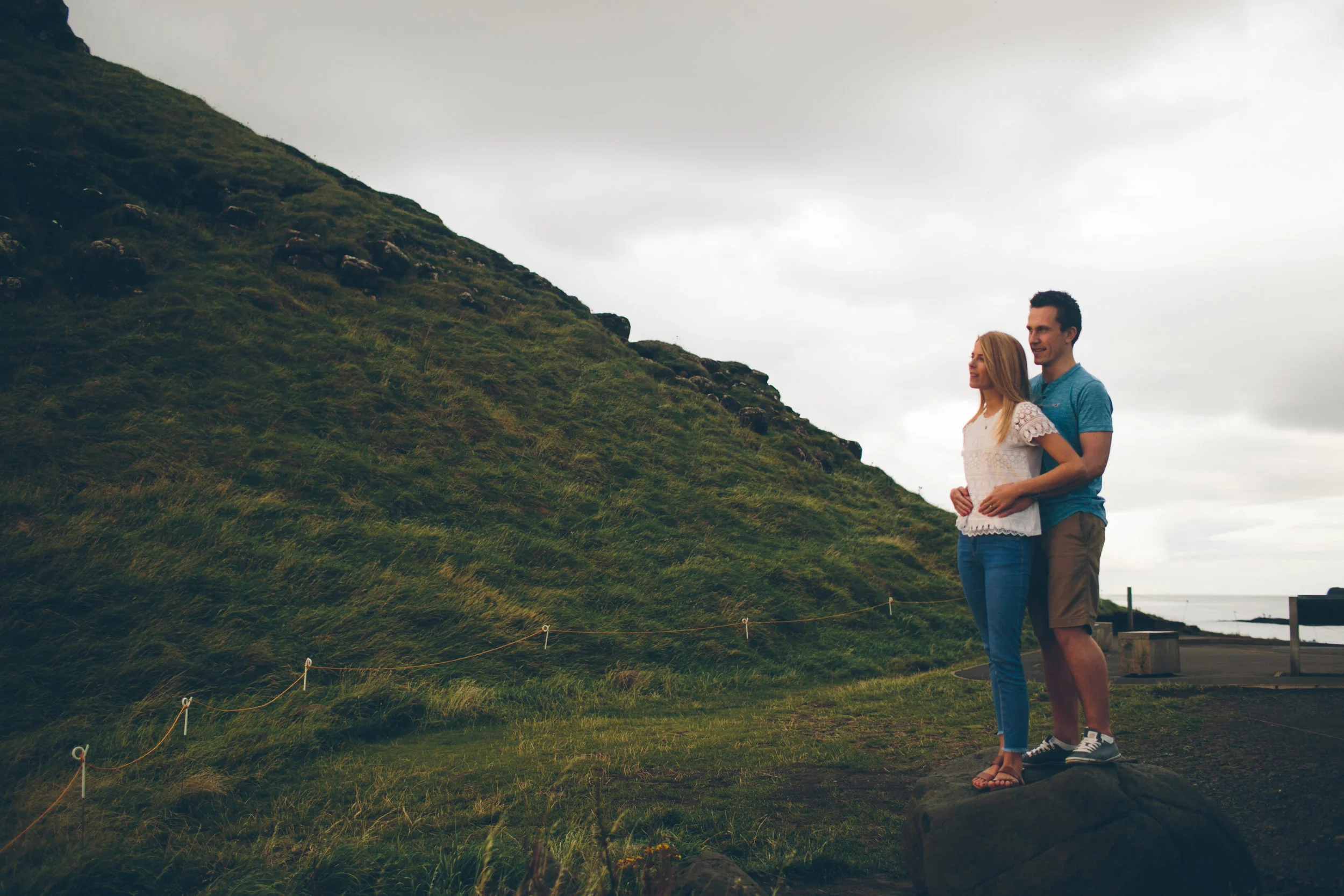 A couple standing on a rock, embracing, by a grassy hill with a cloudy sky in the background.