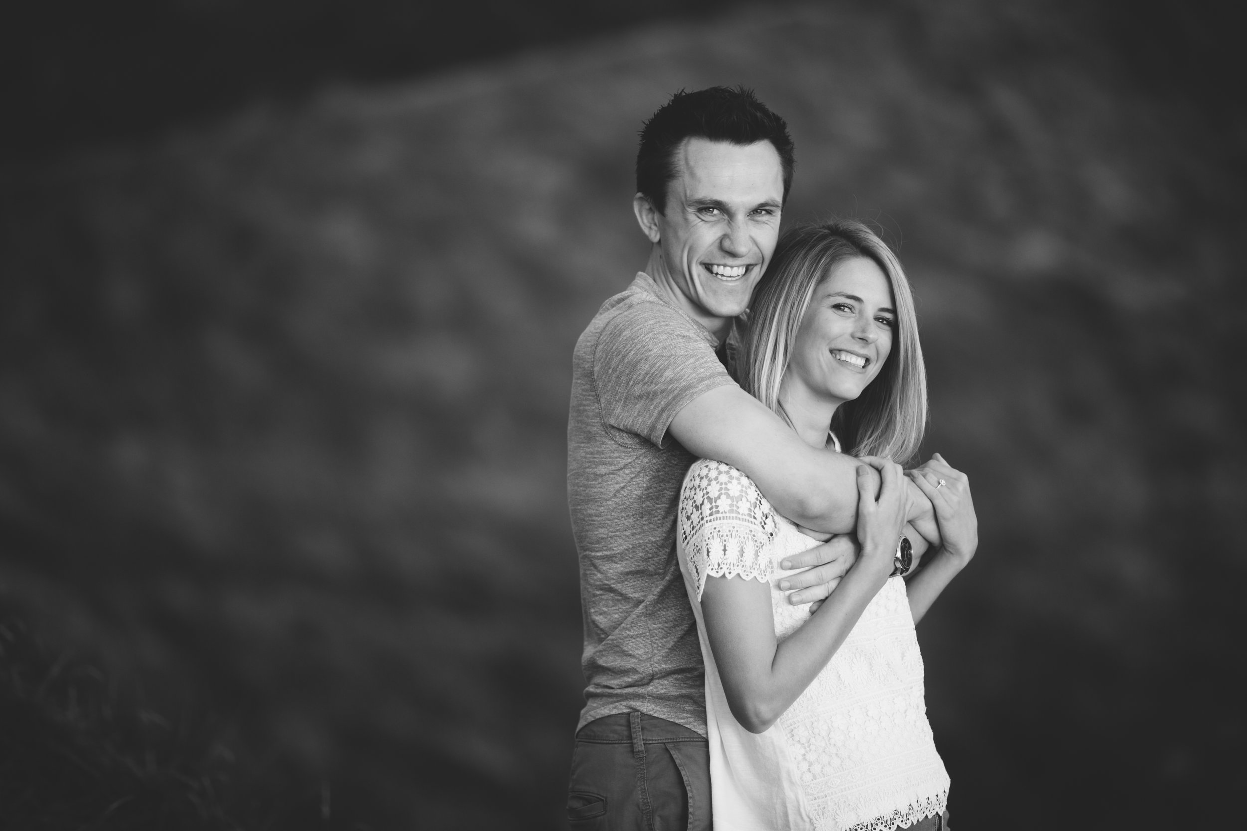 A smiling couple standing outdoors, the man hugging the woman from behind, both looking at the camera.